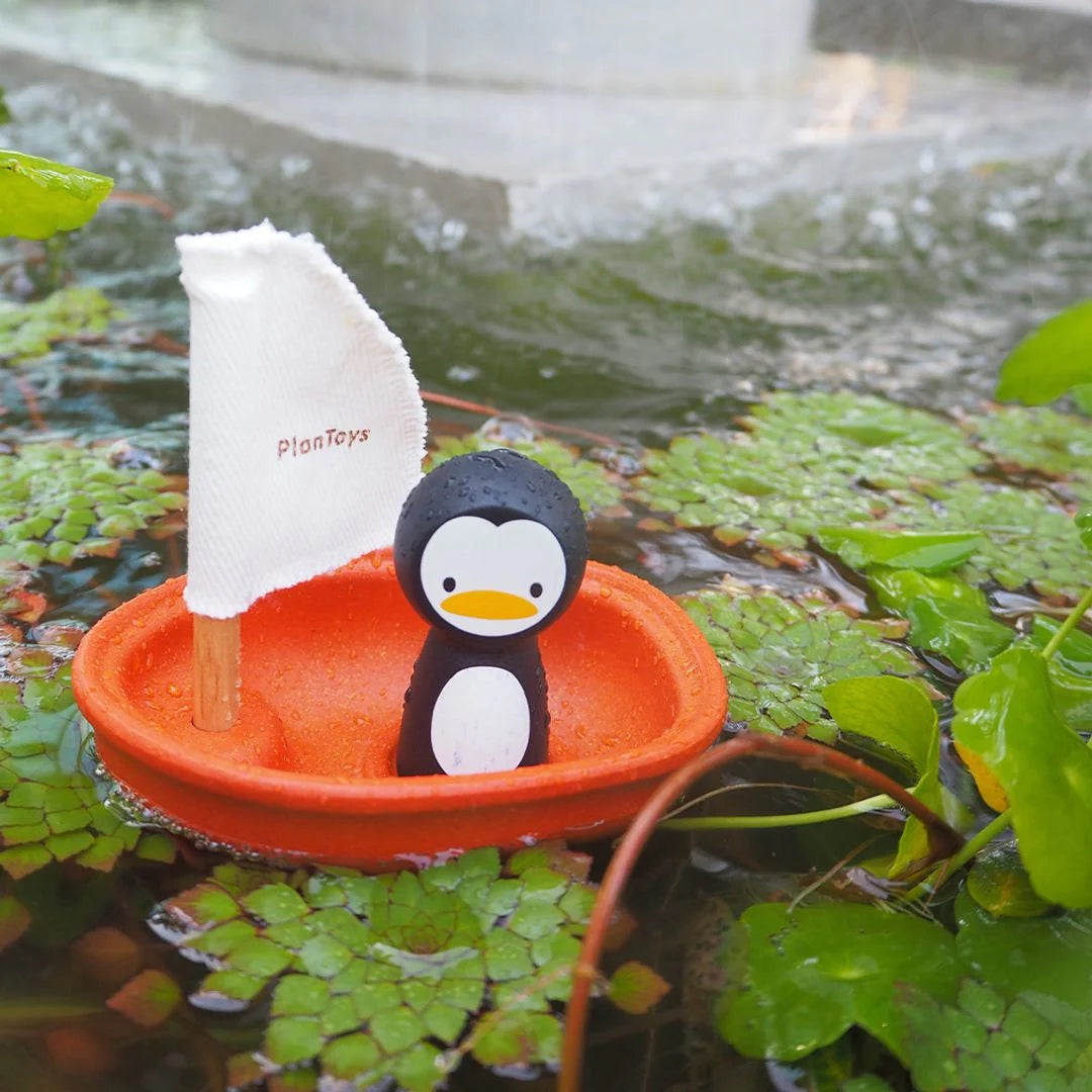 Penguin toy in an orange boat with a white sail on water surrounded by green plants