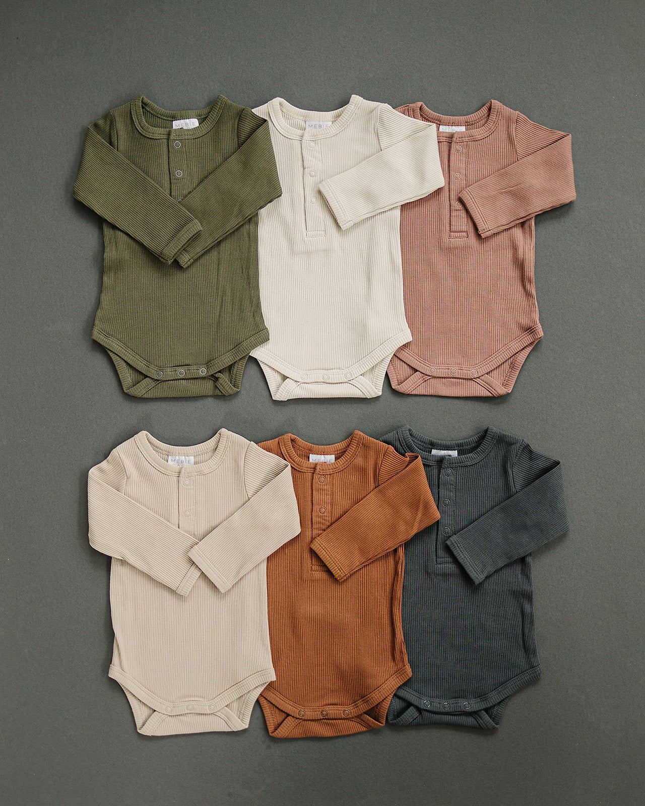 Set of six baby bodysuits in green, beige, pink, gray, brown, and blue on a dark gray background.
