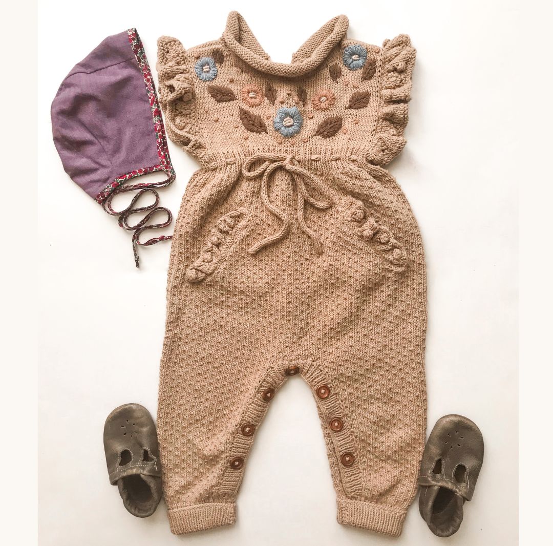 Knitted baby romper with floral patterns, purple bonnet, and brown shoes on a white background