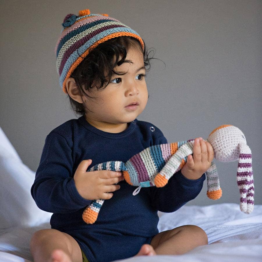 Child wearing a colorful striped hat holding a striped bunny toy on a bed