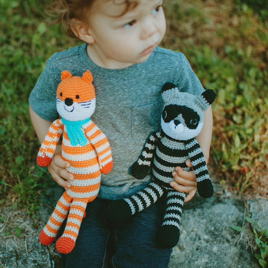 Child holding two crocheted animals, one orange and white striped fox and one black and grey striped raccoon, while sitting outdoors.