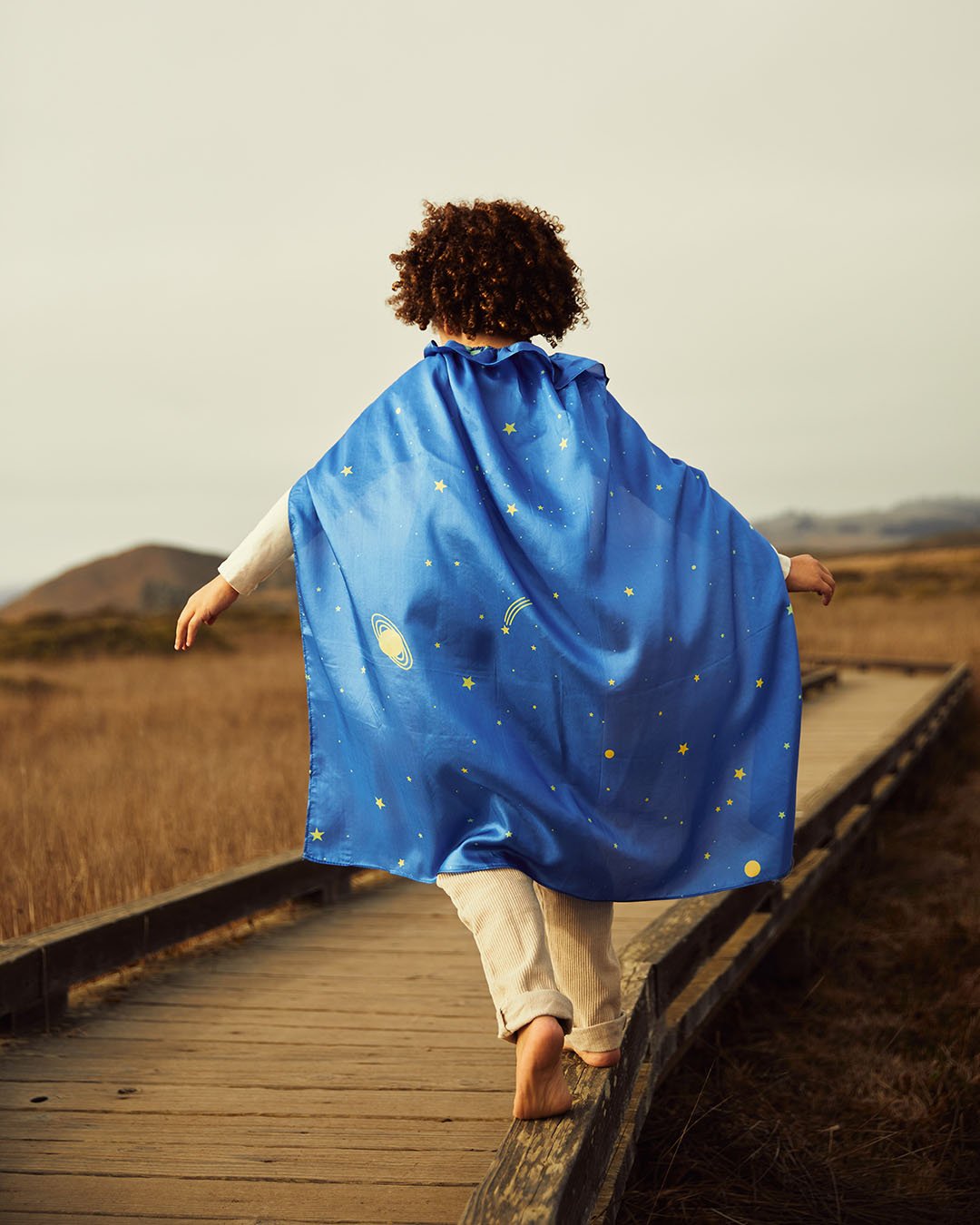 child walking on a boardwalk in a field wearing starry night play cape