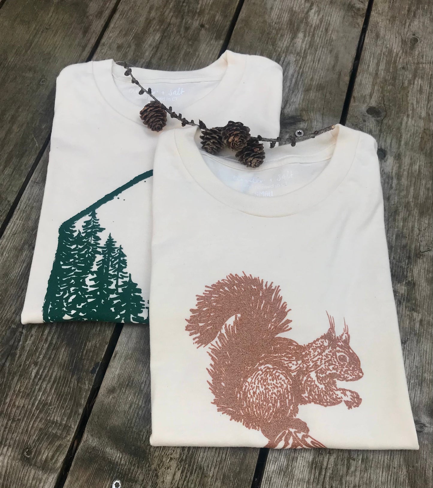 a pnw trees tee and a squirrel tee stacked on a wooden table with decorative pinecones