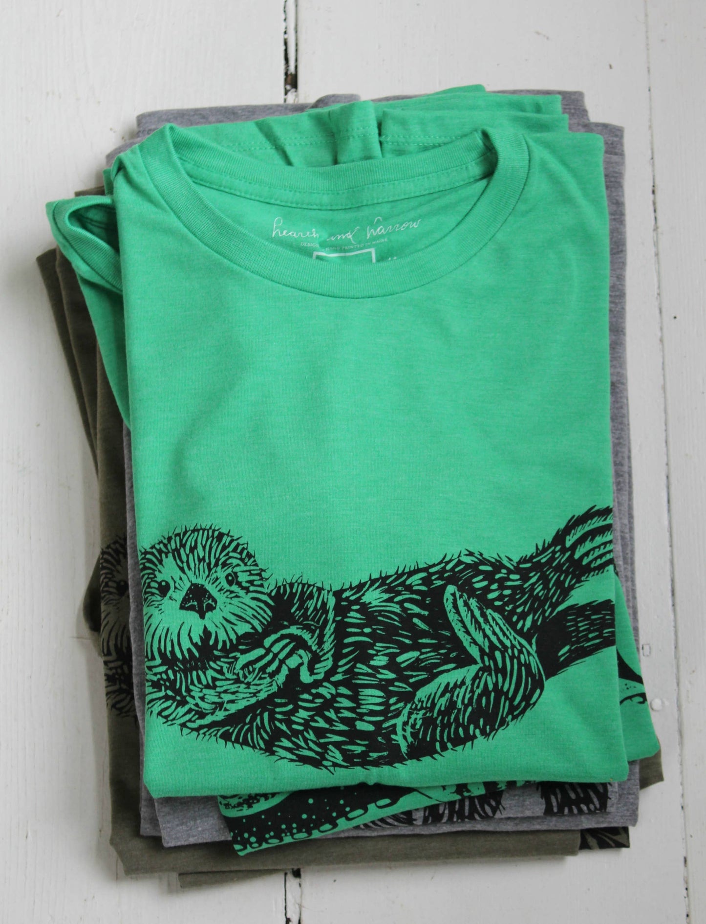 Stack of green t-shirts with a black otter design on a white wooden surface