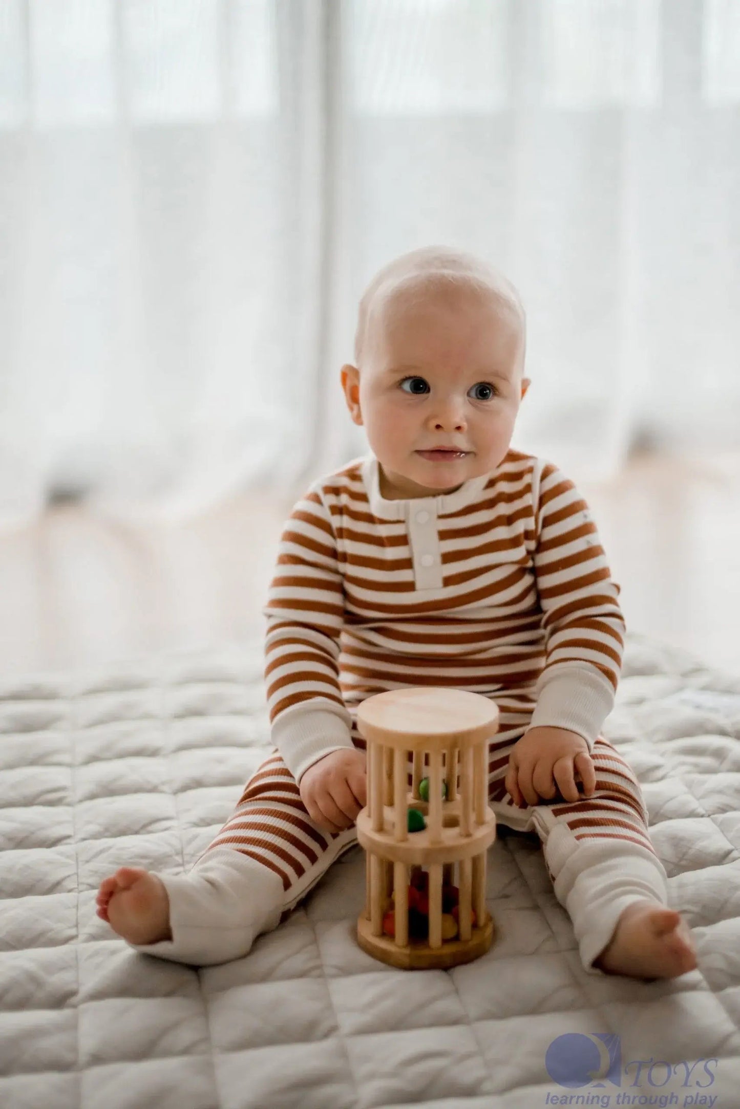 a baby sitting on a floor play mat wearing a striped romper playing with a wooden mini rain maker