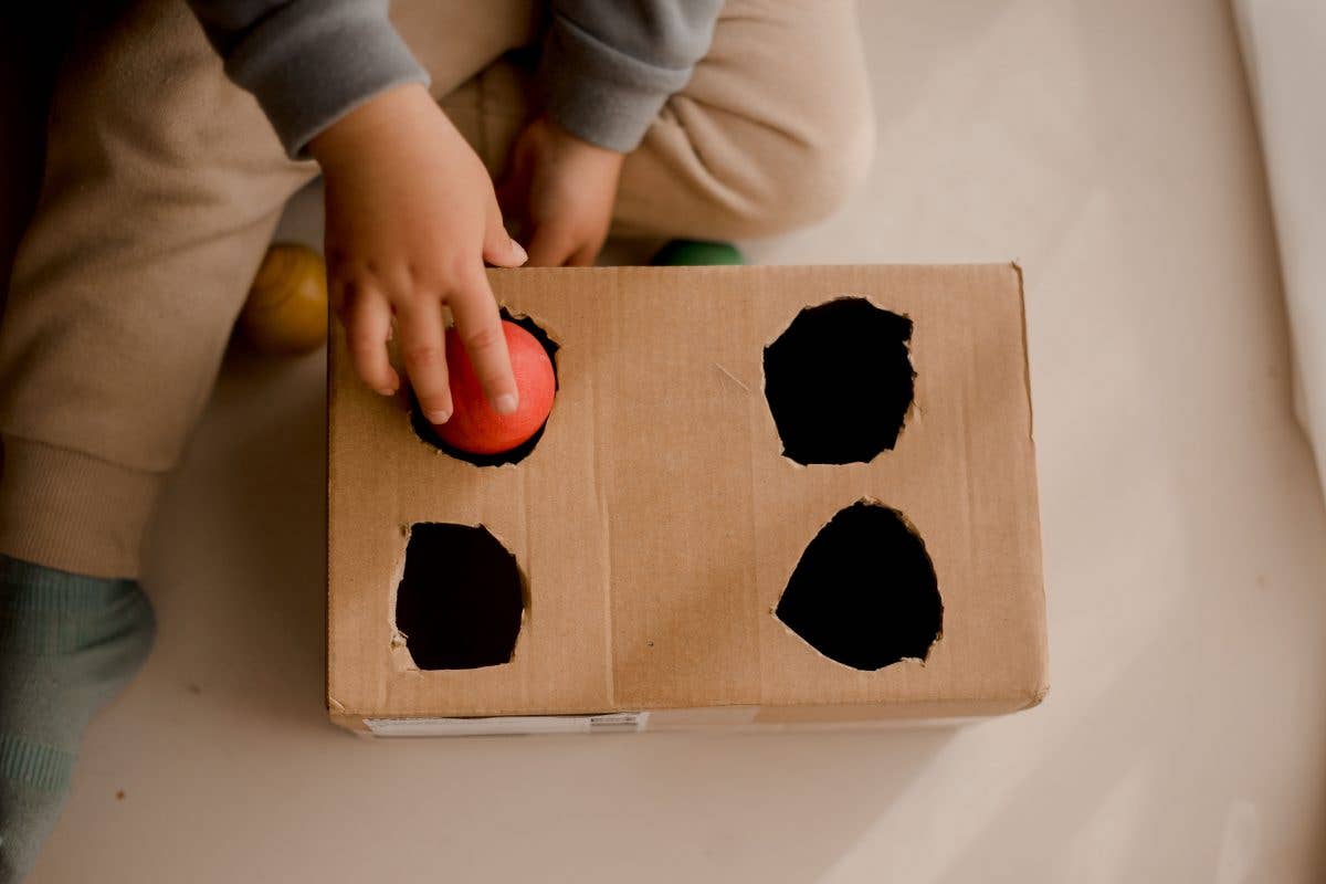 Child pushing a red wooden ball into a cardboard box with holes cut out on the top on a beige surface