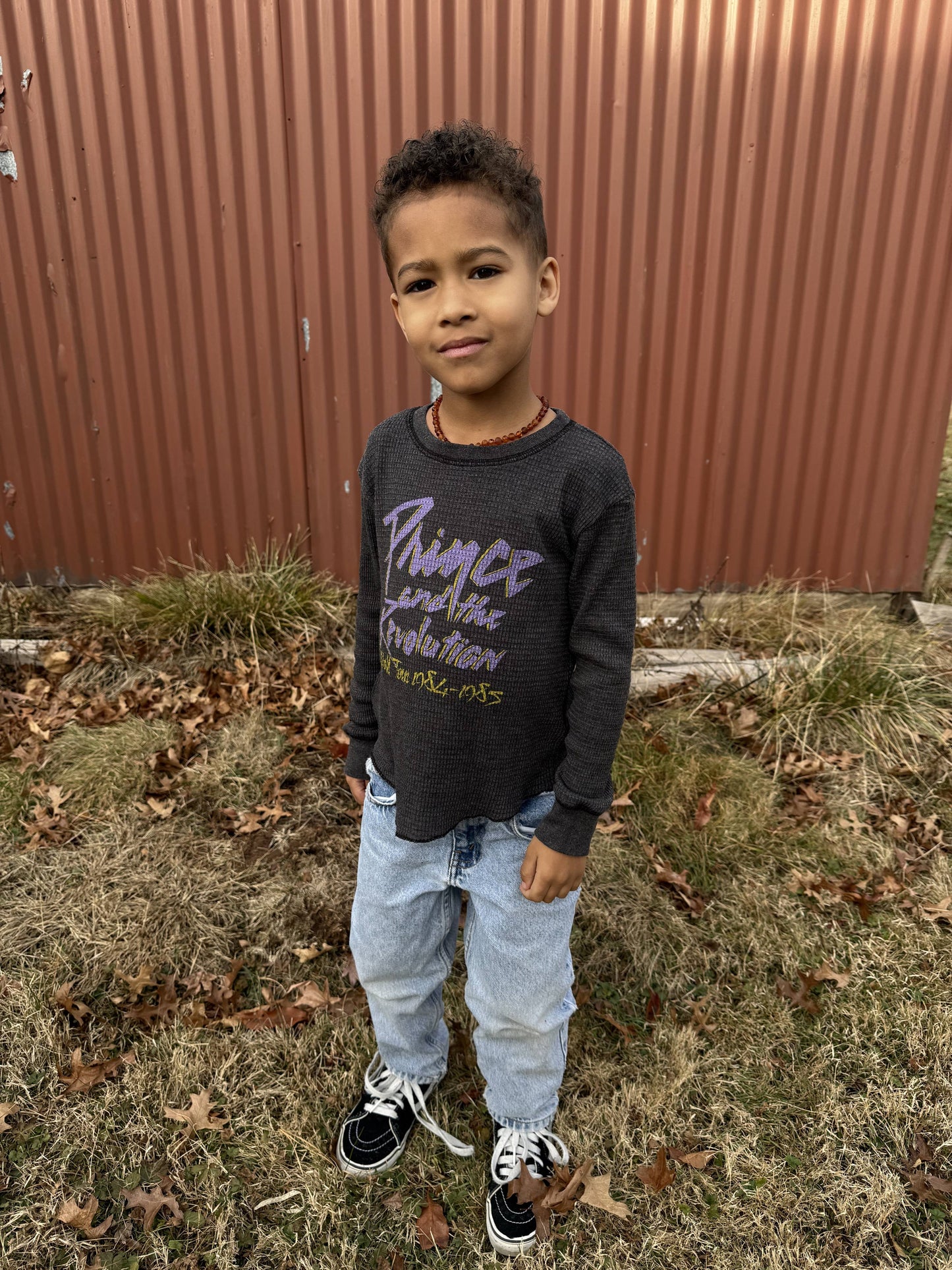 Child wearing a dark gray thermal tee with text, standing in front of a red metal fence.