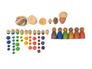 A series of small elements arranged on a plain surface. On top, three round pieces of wood, a small acorn, a pinecone, and a large acorn are arranged in a row. Below and to the left, small acorns and beads of various sizes are arranged in columns based on