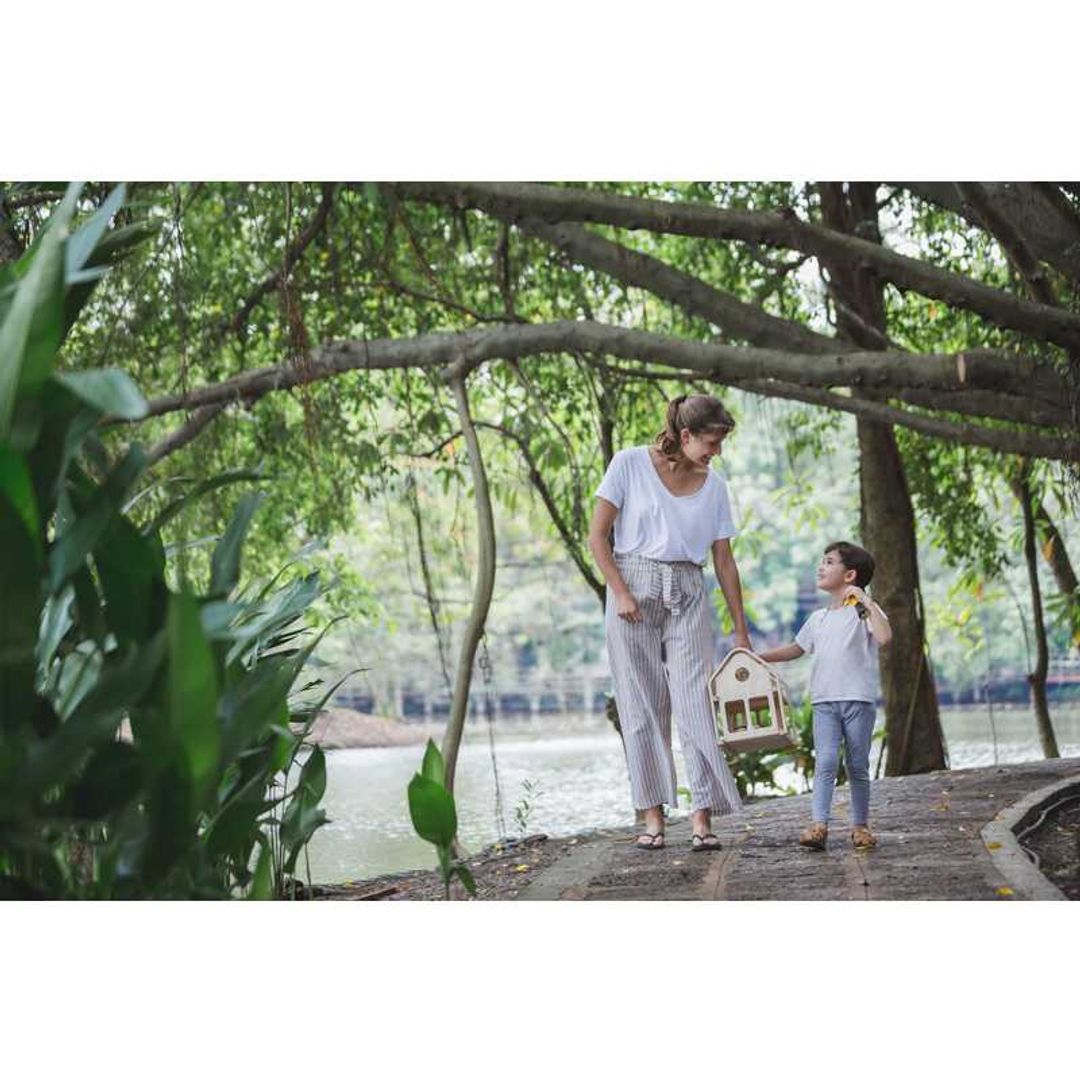 Adult and child carrying Slide N Go Doll house on a path with tree's behind them by Plan Toys