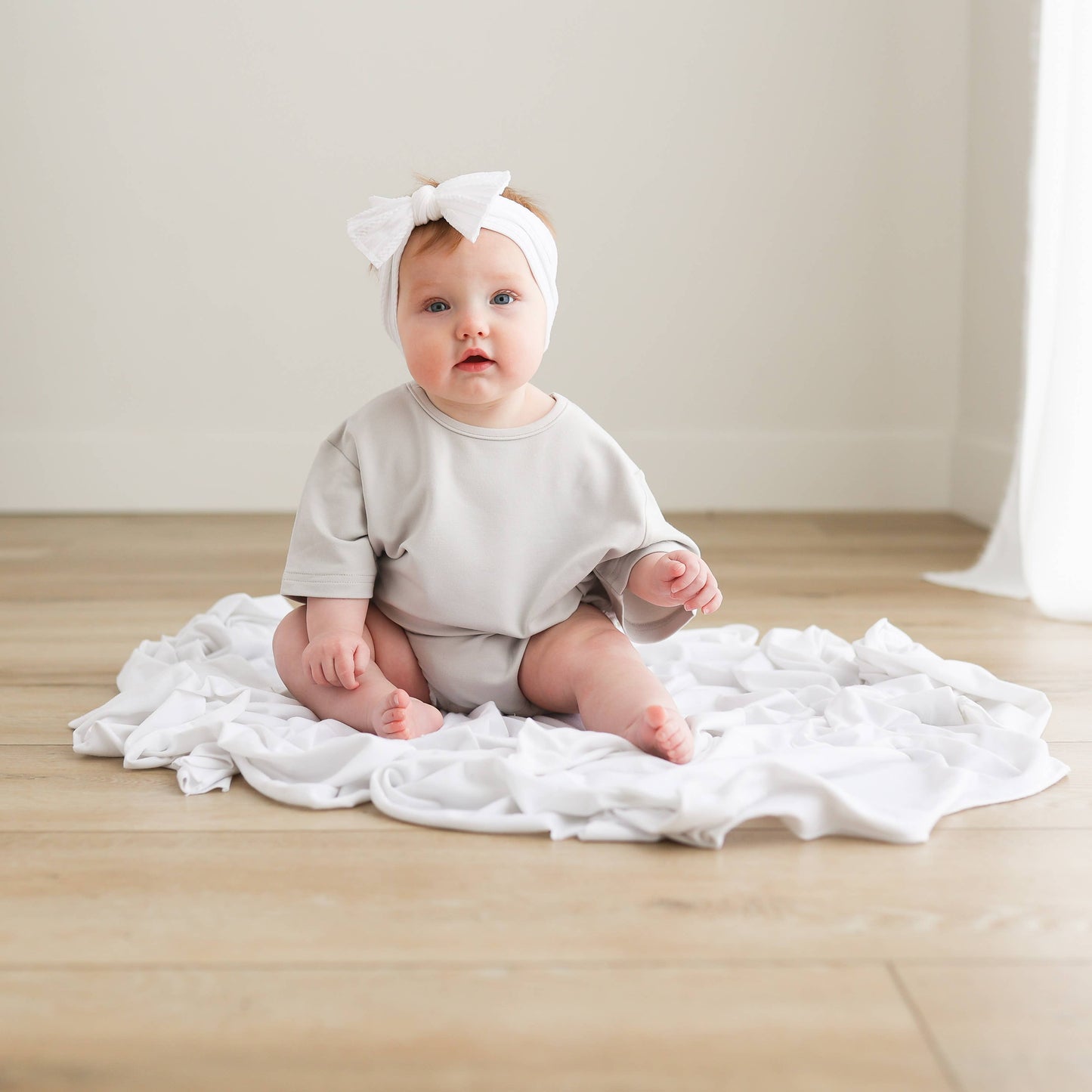 Baby sitting on a white blanket wearing a light gray outfit and white headband.