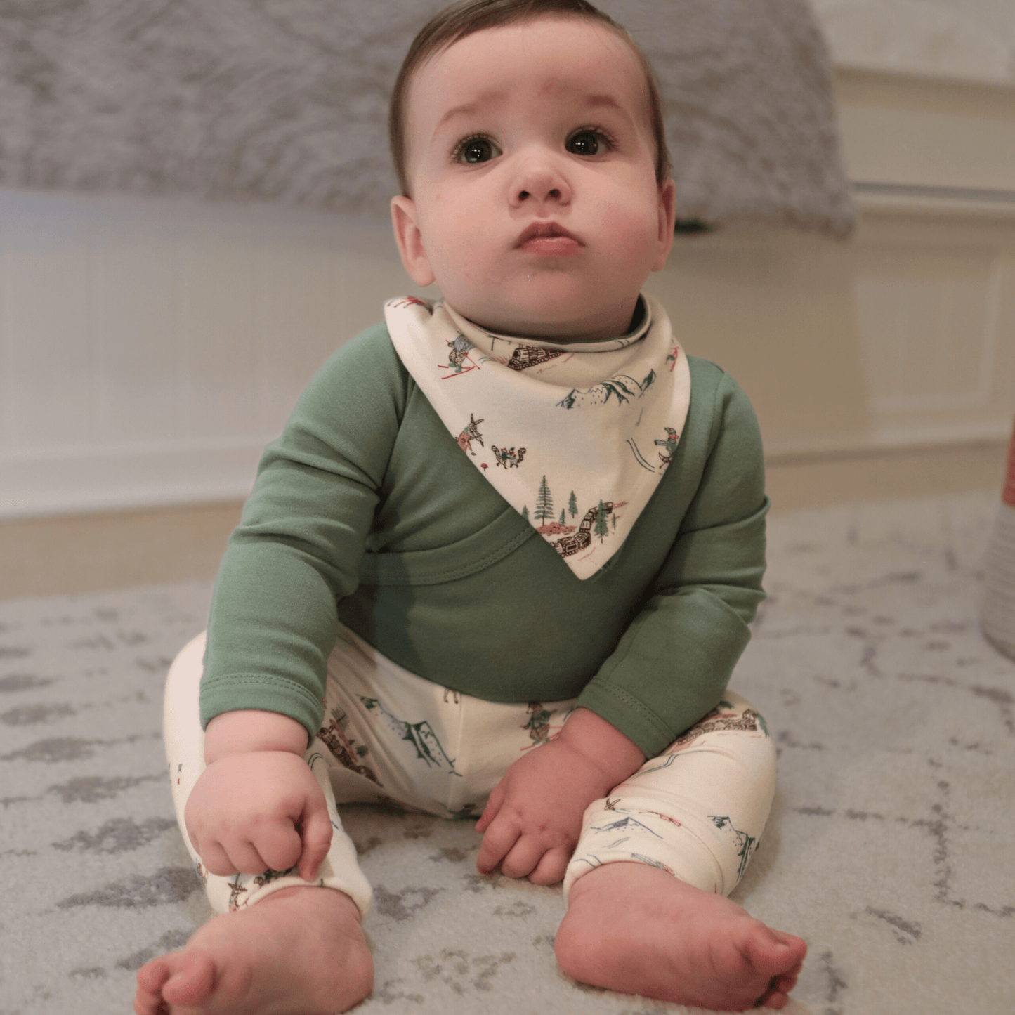 Baby sitting on a carpeted floor wearing a green long-sleeve shirt and patterned pants.