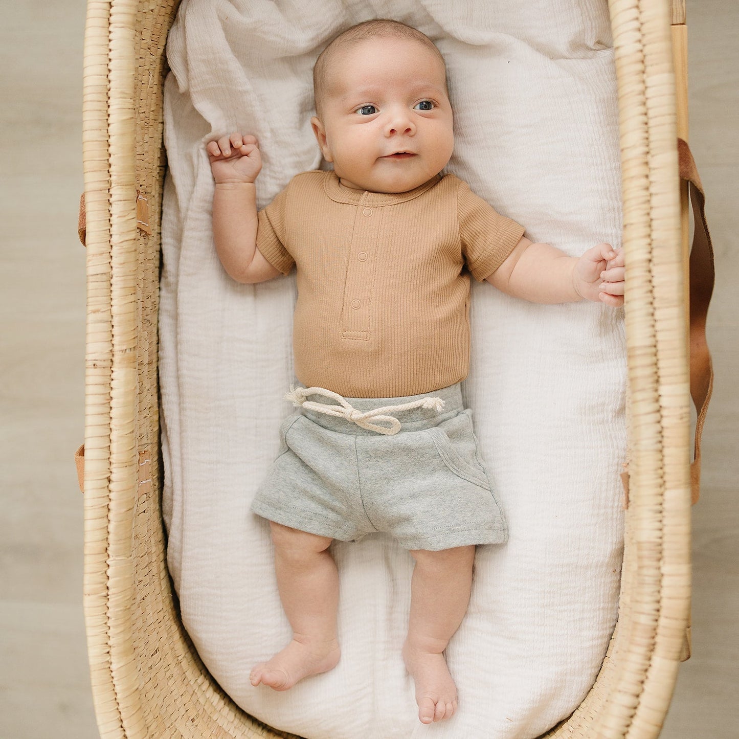 Baby lying in a moses basket wearing a brown shirt and gray shorts.