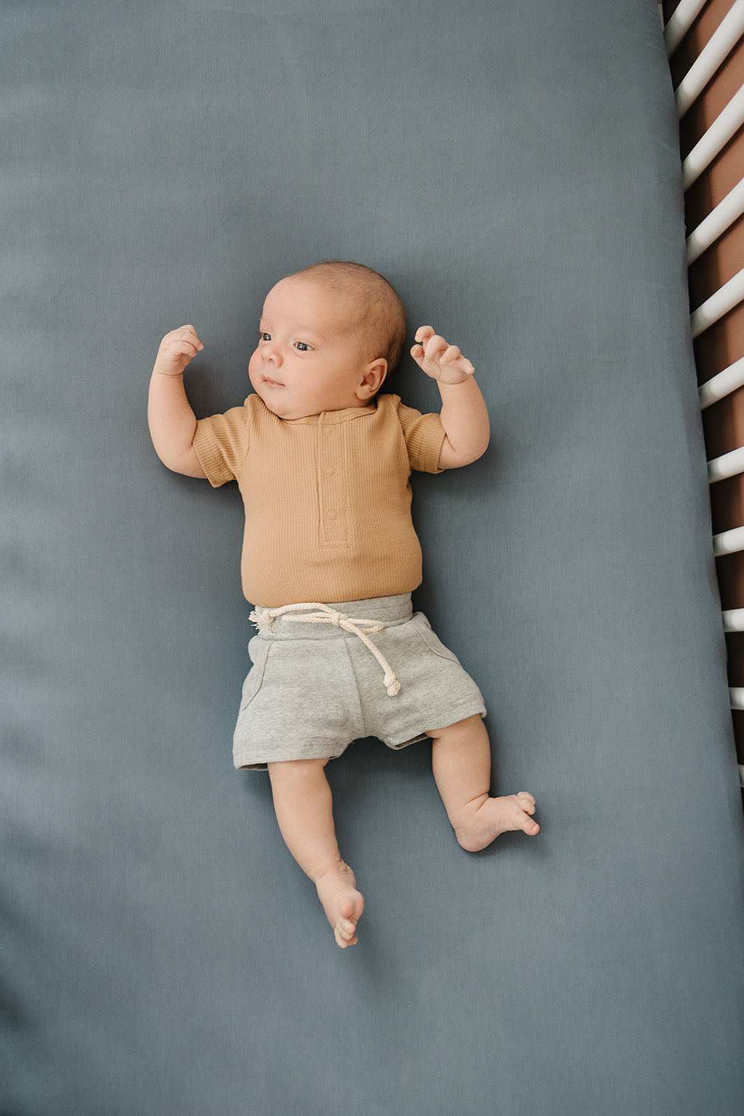 Baby lying on a gray surface wearing a brown shirt and gray shorts.