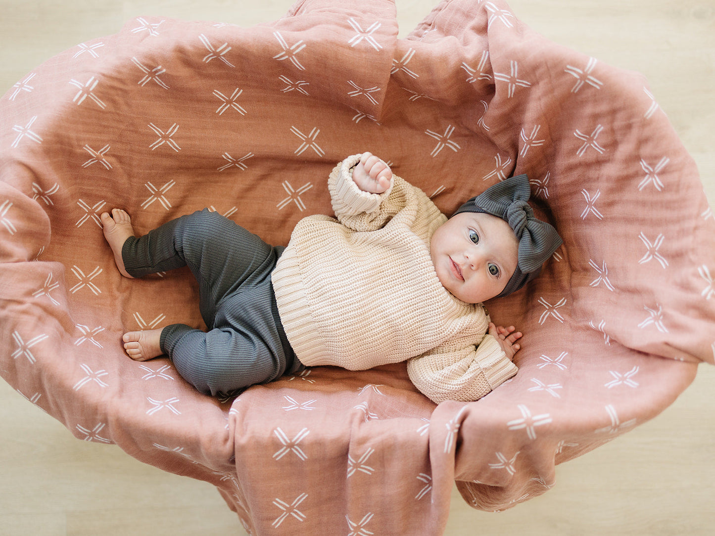 Baby lying in a pink patterned crib wearing a beige sweater and gray pants.