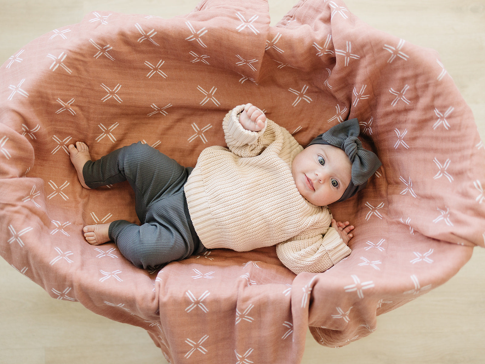 Baby lying in a pink patterned crib wearing a beige sweater and gray pants.