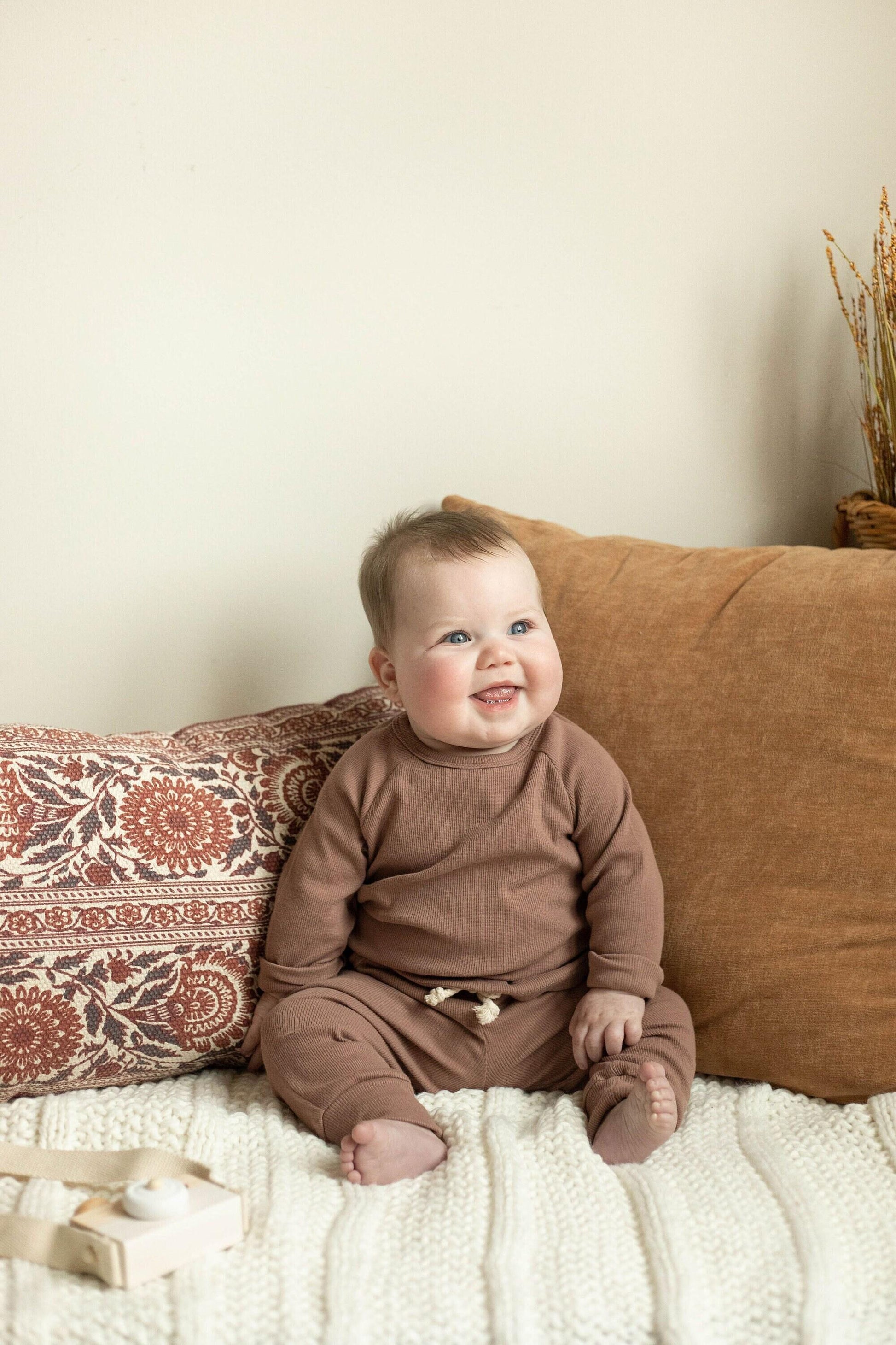 Baby in a brown outfit sitting on a couch with pillows and a neutral background