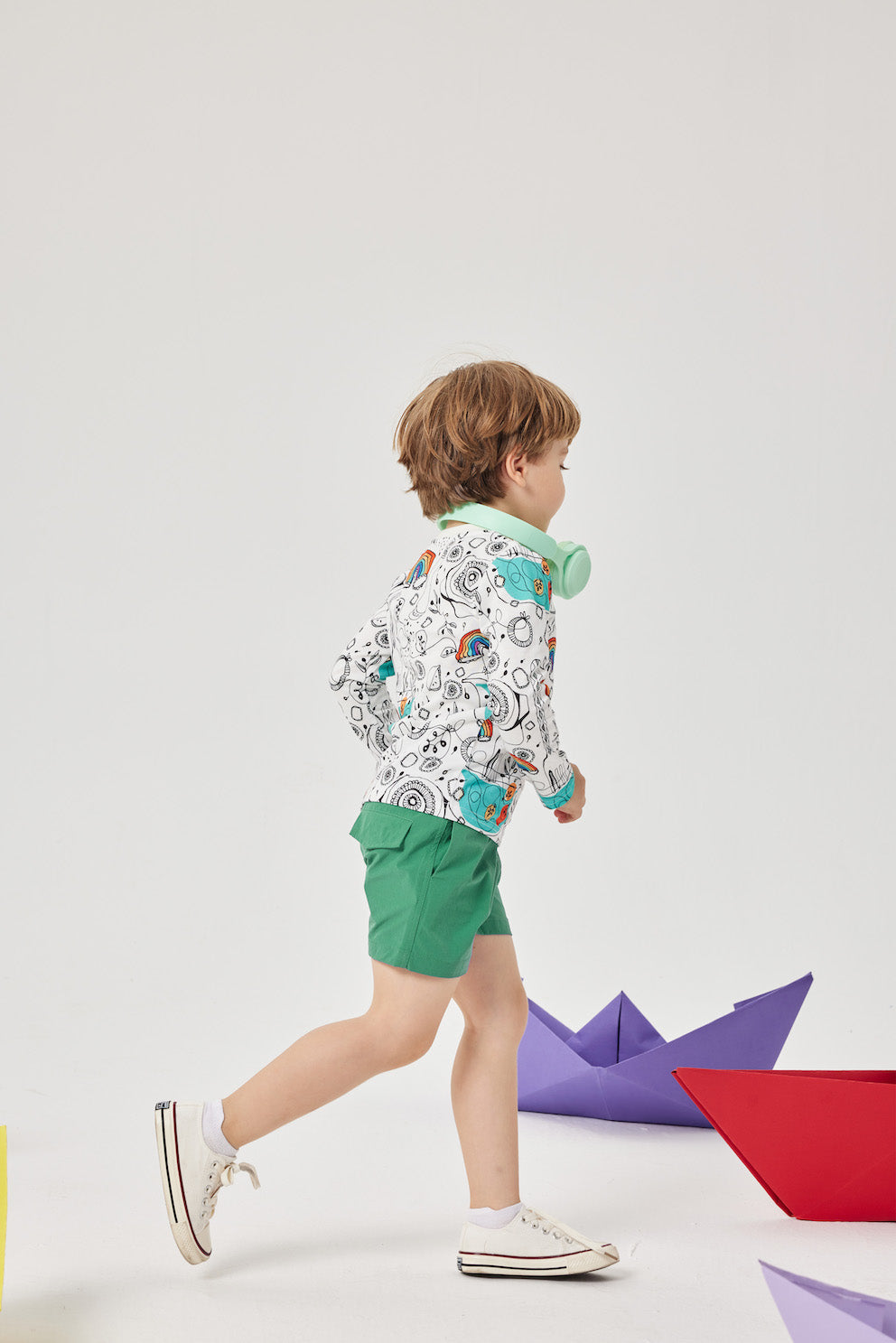 Child wearing a patterned jacket and green shorts standing on a white background with colorful paper boats.
