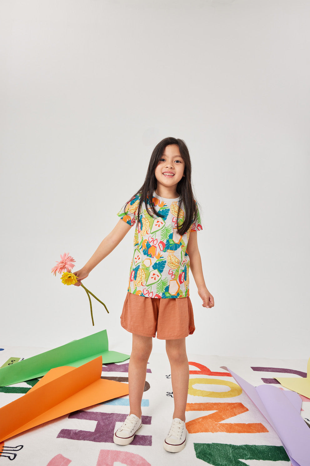 Young girl in a floral top and orange skirt standing on a colorful rug with a white background