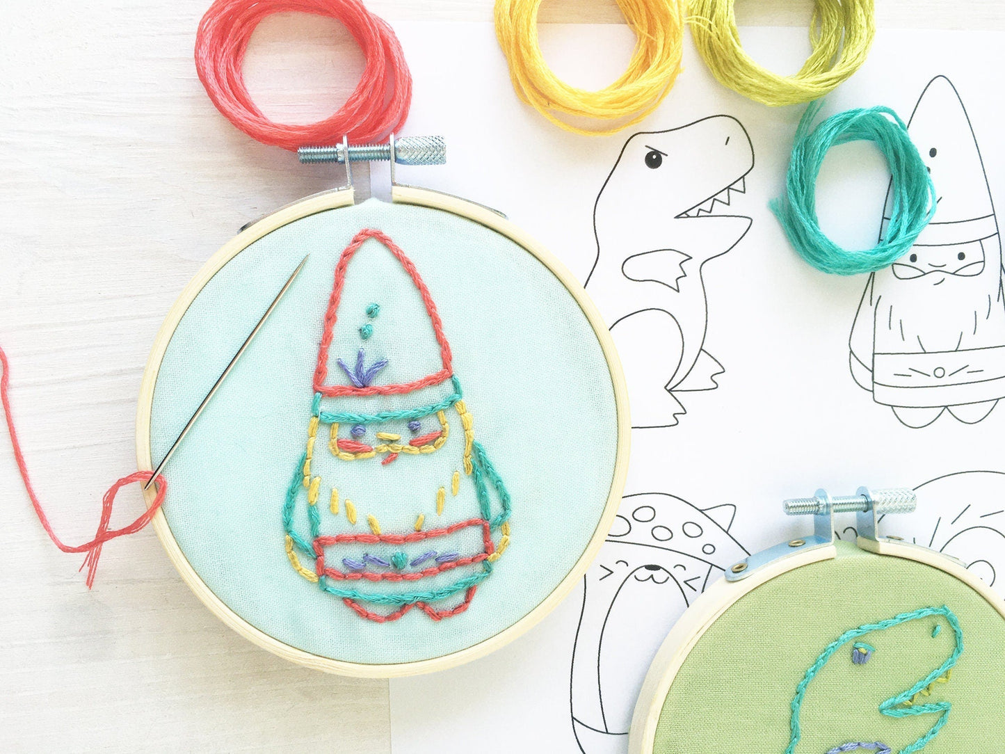 Embroidery hoop with colorful embroidered design on a light blue background, surrounded by yarn and dinosaur outlines.