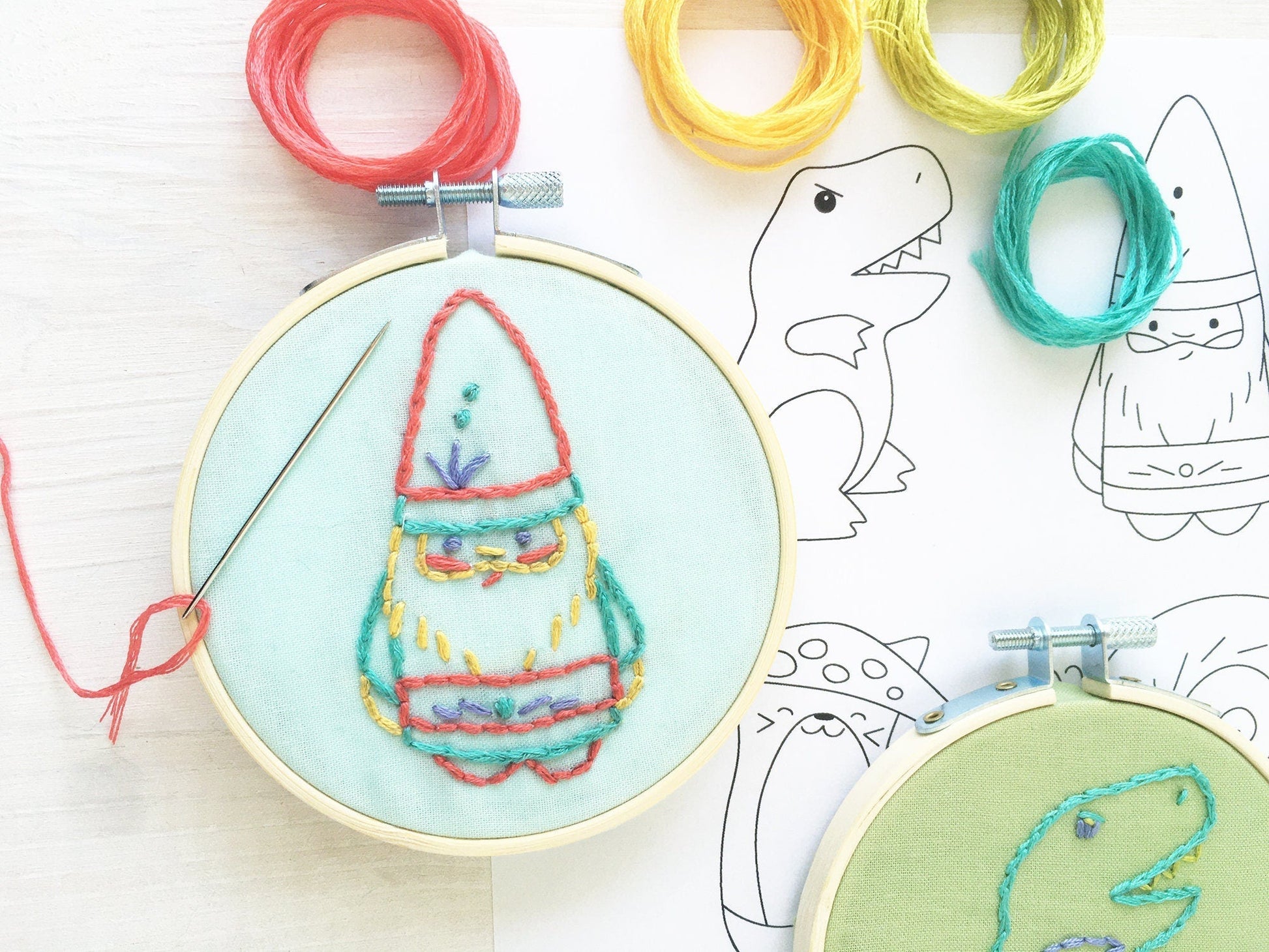 Embroidery hoop with colorful embroidered design on a light blue background, surrounded by yarn and dinosaur outlines.