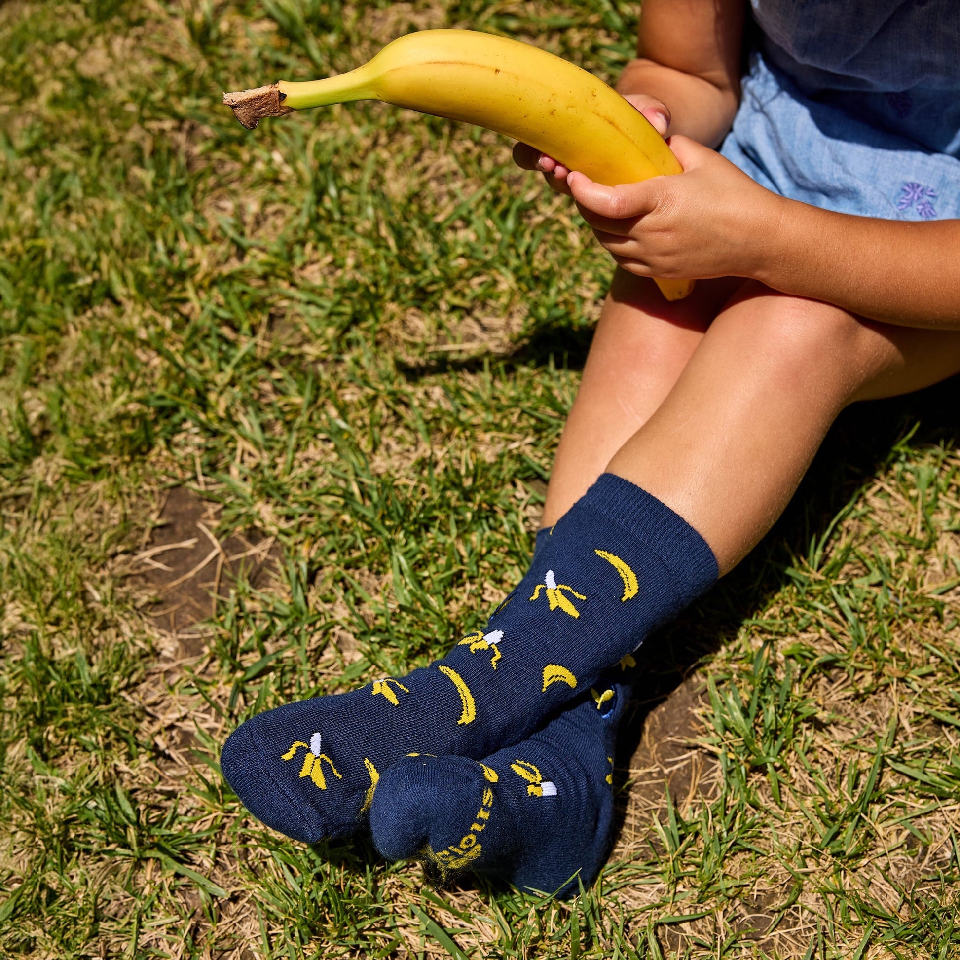 Person wearing navy blue socks with banana pattern sitting on grass holding a banana