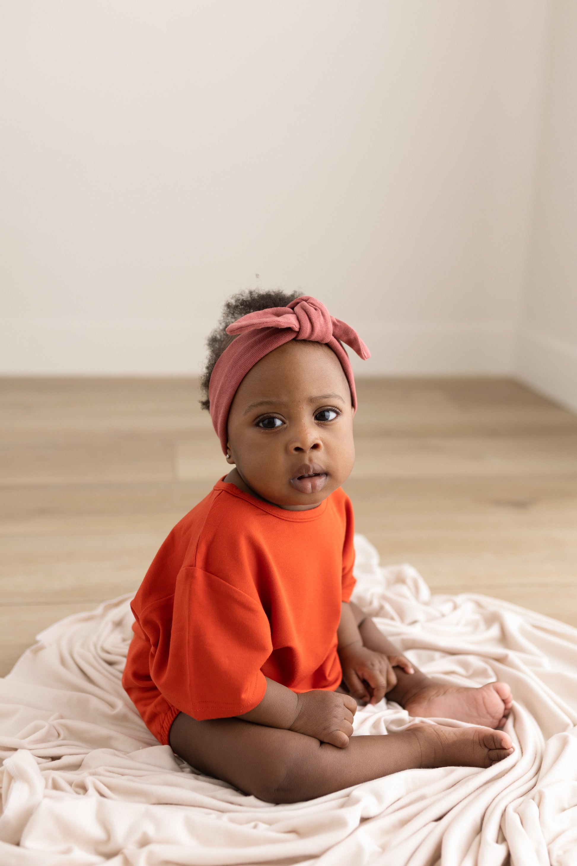 Baby wearing an orange shirt and pink headband sitting on a white blanket.