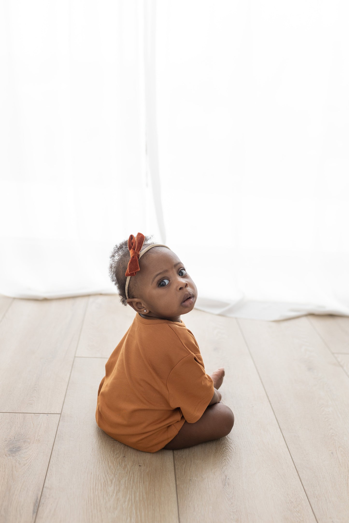 Baby sitting on a light wooden floor wearing an orange outfit with a red headband.
