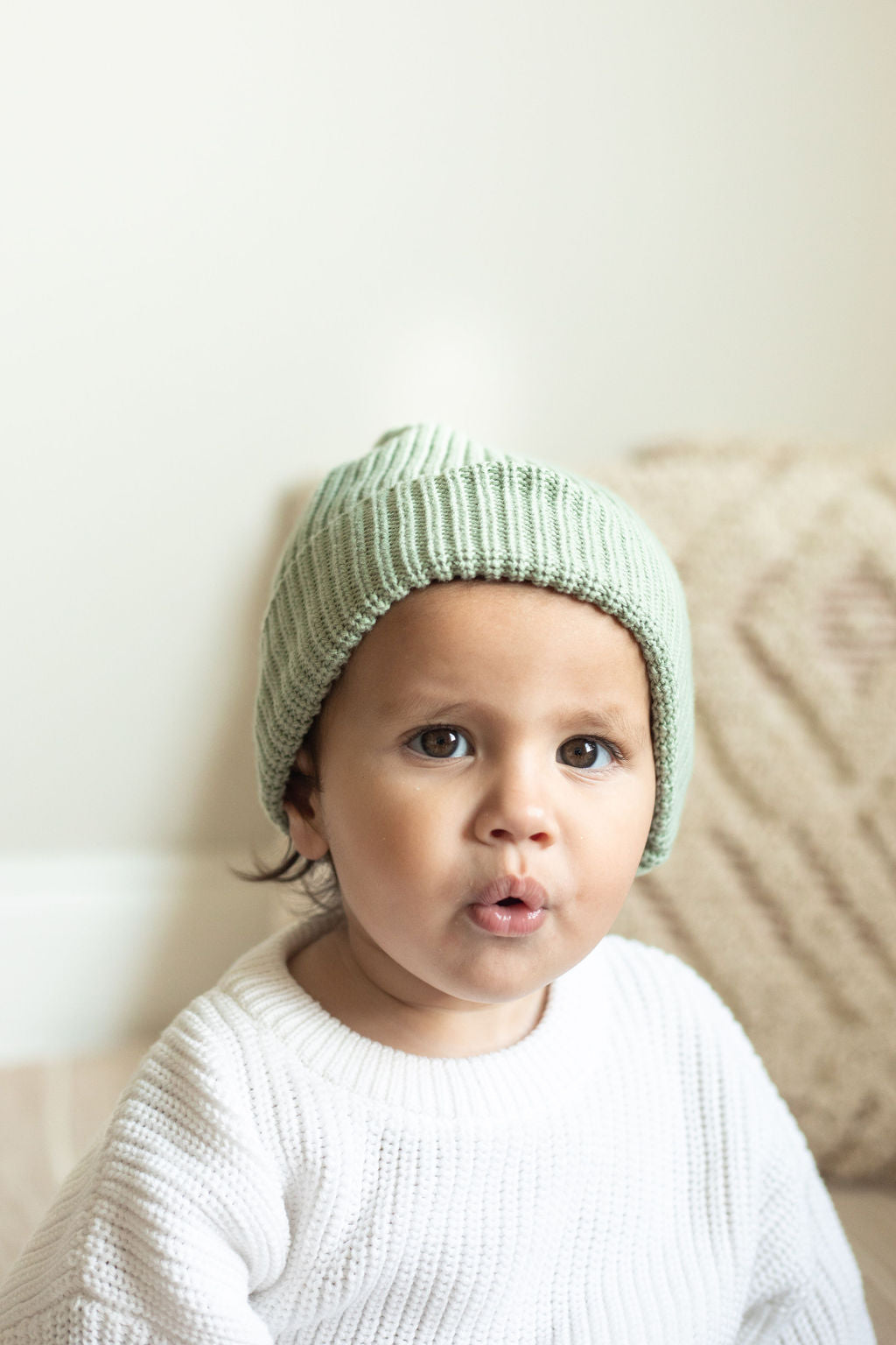 Baby wearing a green knit hat sitting on a beige couch.