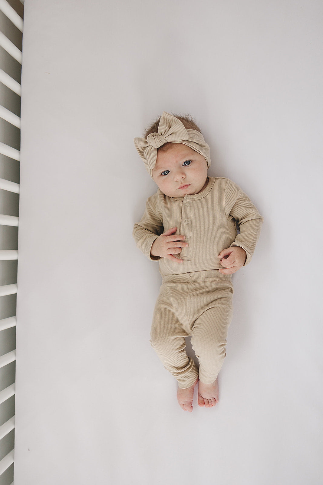 Baby in a beige outfit lying on a white surface with a white crib in the background