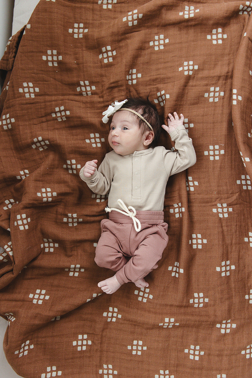 Baby lying on a brown patterned blanket wearing a beige top and pink pants.