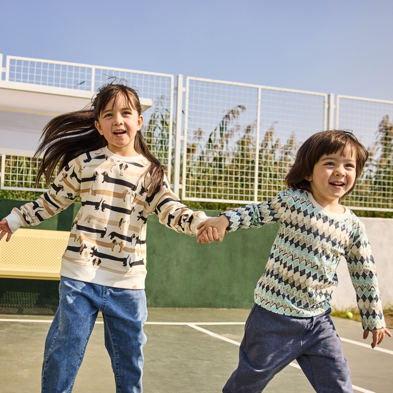 Two children holding hands on a tennis court with a clear blue sky.
