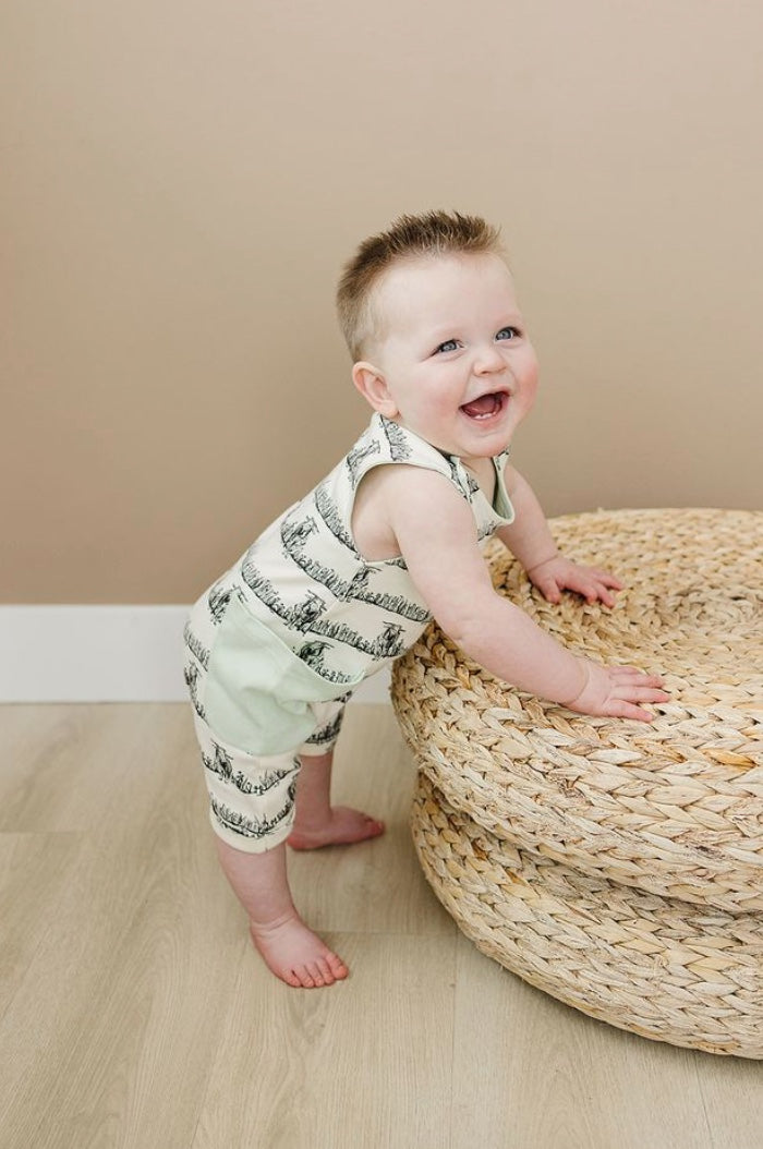 Baby in a striped romper standing on a woven mat against a beige wall.