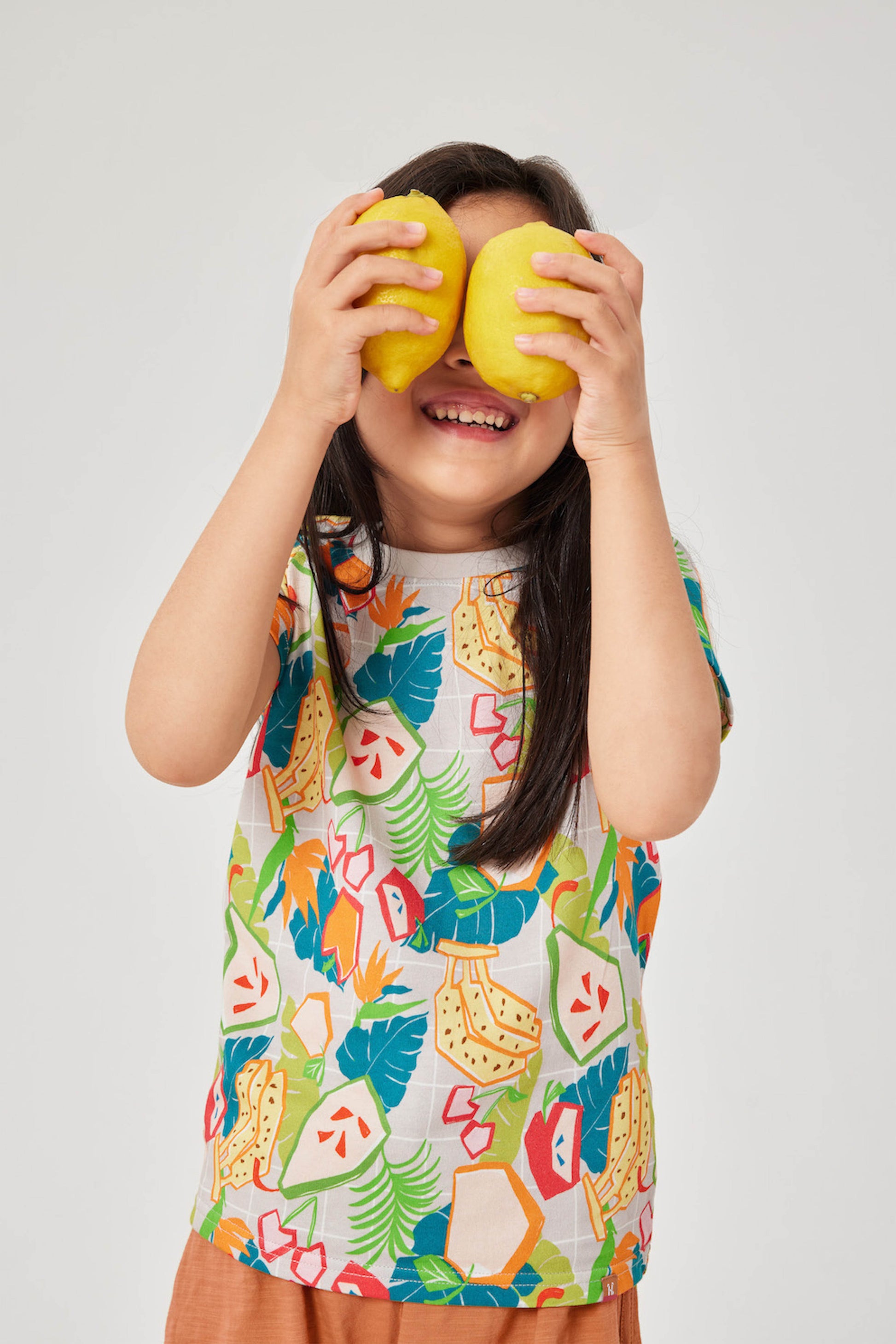 Child wearing a colorful shirt with fruit patterns, holding two yellow fruits in front of their face on a white background.