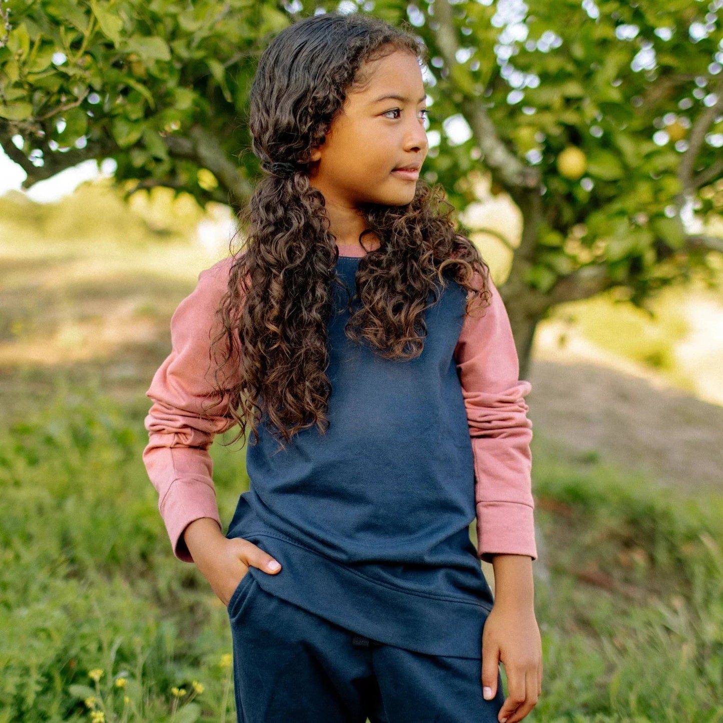 Young girl in a blue dress and pink shirt standing outdoors with trees in the background