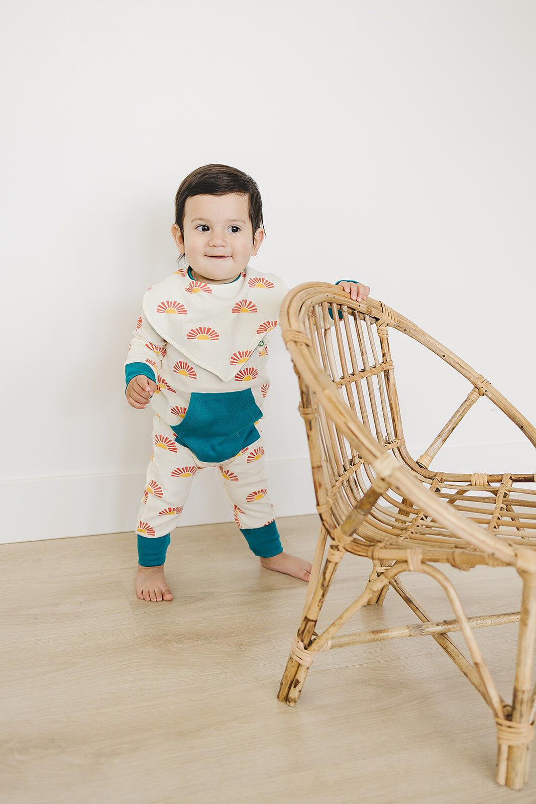 Child wearing a patterned outfit standing next to a wicker chair on a light wooden floor.
