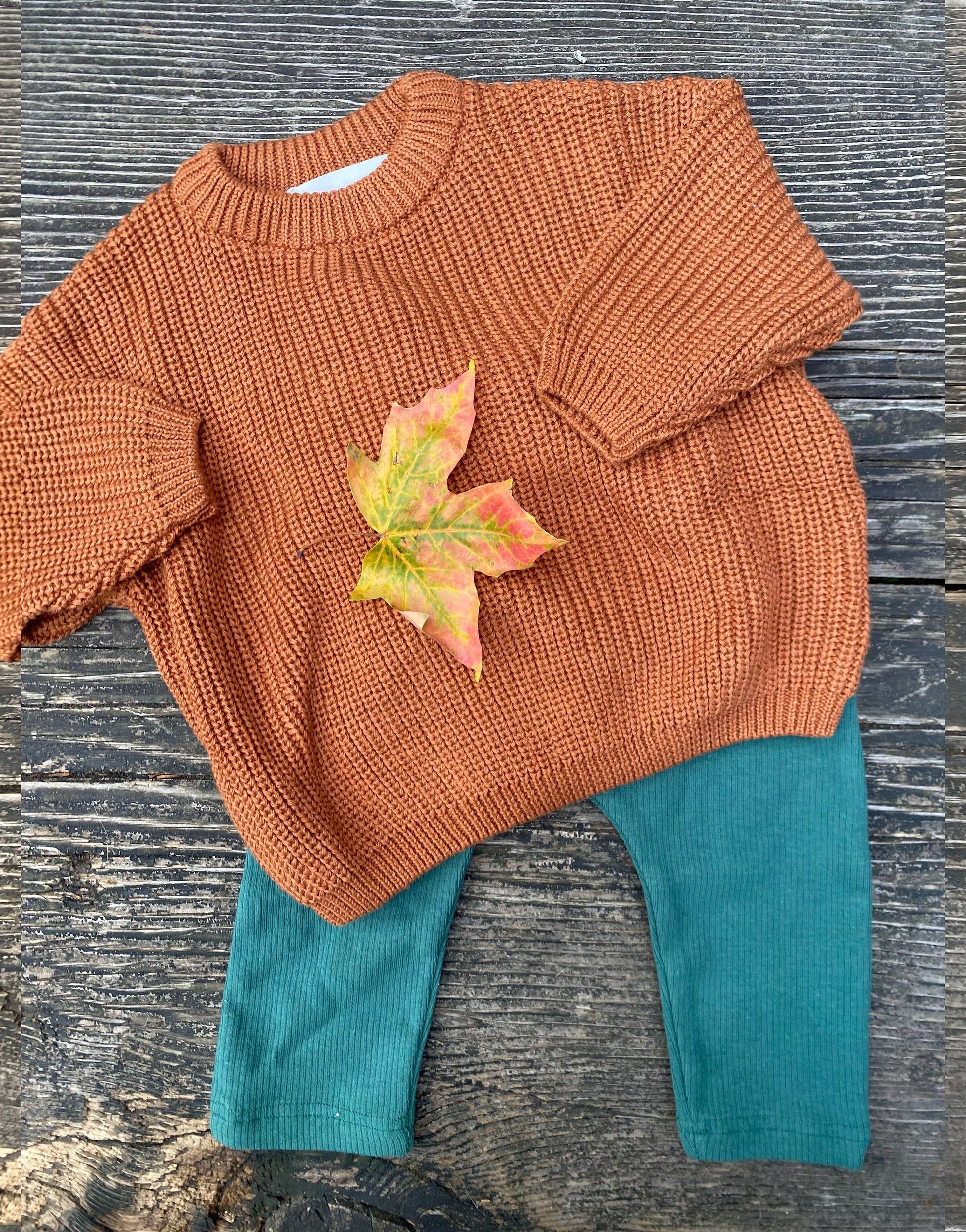 Clay  knitted sweater with a colorful leaf on top of teal pants on a textured surface