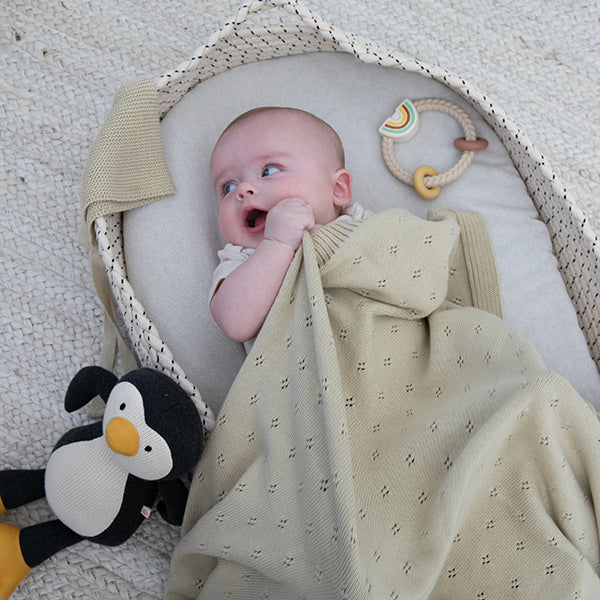 Baby wrapped in a beige blanket with a penguin toy next to it, lying on a textured surface.