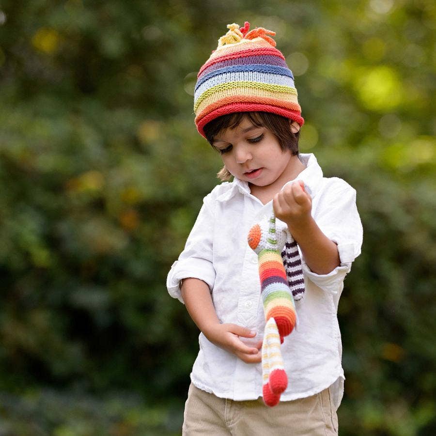 Child wearing a colorful striped hat and holding a matching rainbow striped bunny toy outdoors.