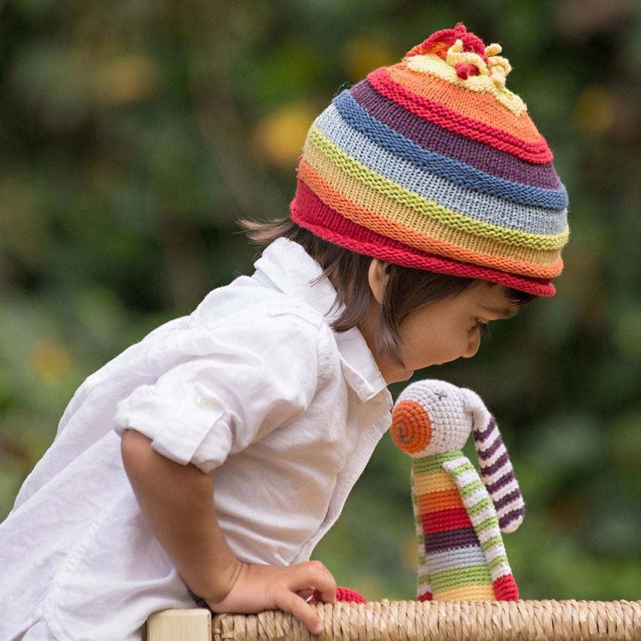 Child wearing a colorful striped hat holding a matching stuffed toy outdoors.