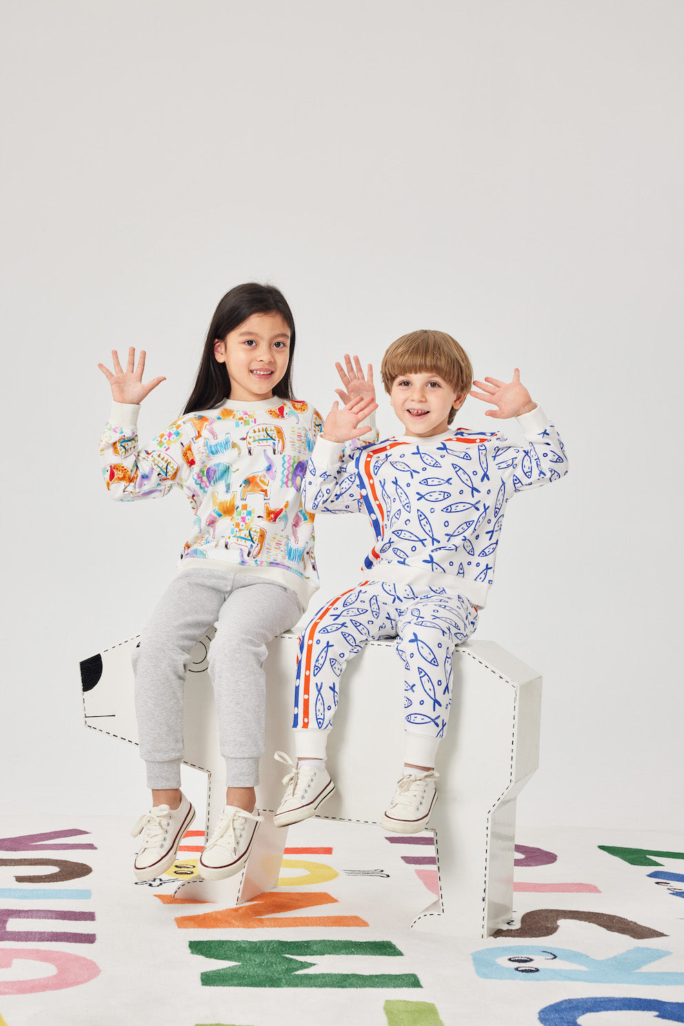 Two children wearing colorful outfits sitting on a chair with a white background.