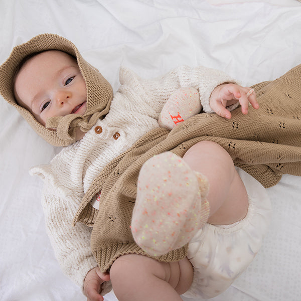 Baby in a knitted outfit with a hood and socks, lying on a white blanket.