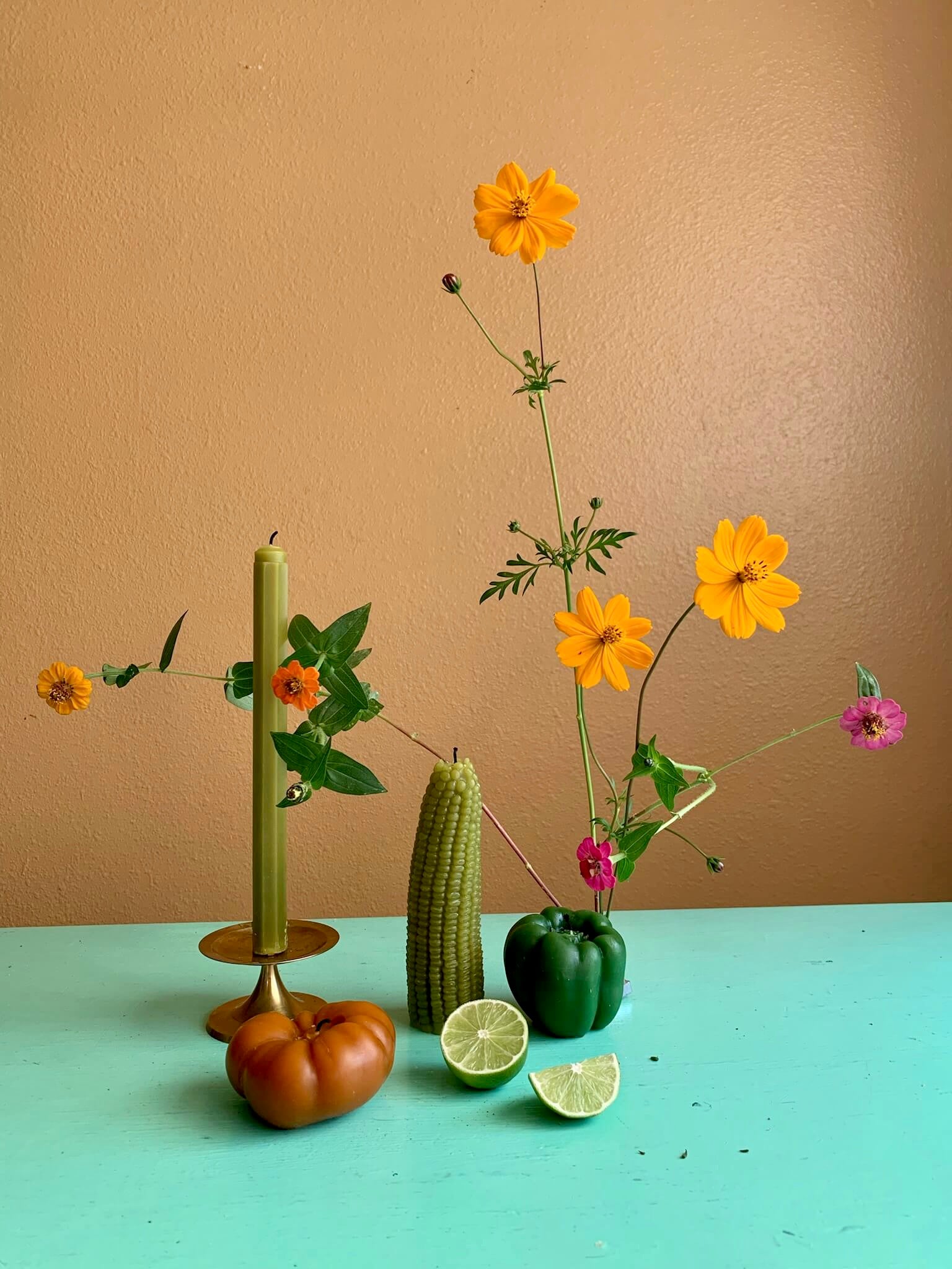 Still life arrangement with flowers, vegetables, and candles on a teal surface against a brown background