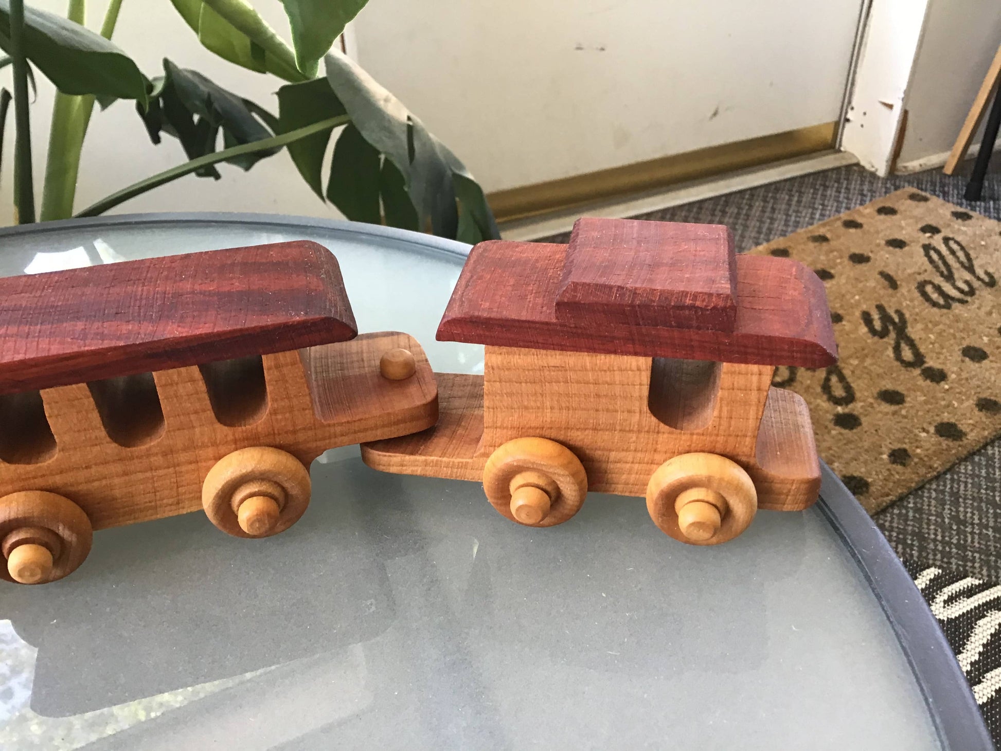 Wooden toy train on a reflective surface with a plant and doormat in the background