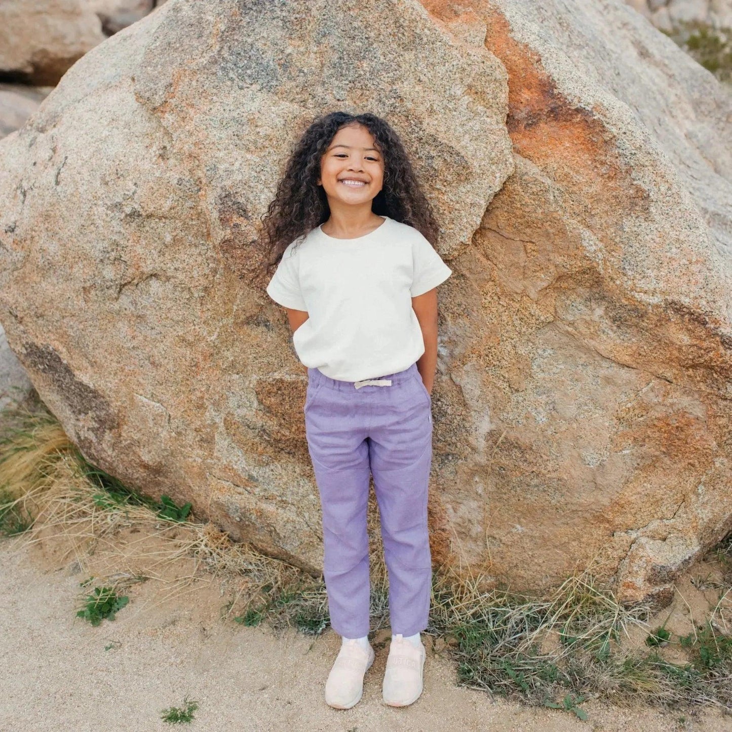 Young girl standing in front of a large rock, wearing a white shirt and purple pants.