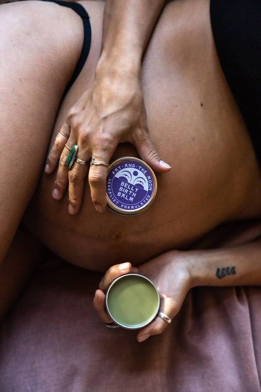 a pregnant person laying on a cloth holding a tin of belly birth balm by Fat and the Moon