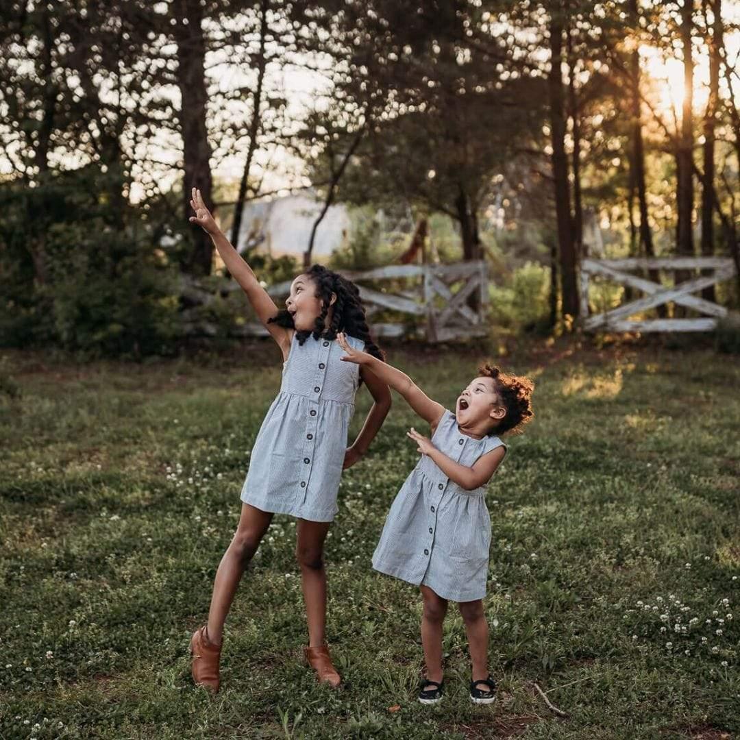 Two young girls in matching dresses playing in a grassy field with trees and a sunset in the background.