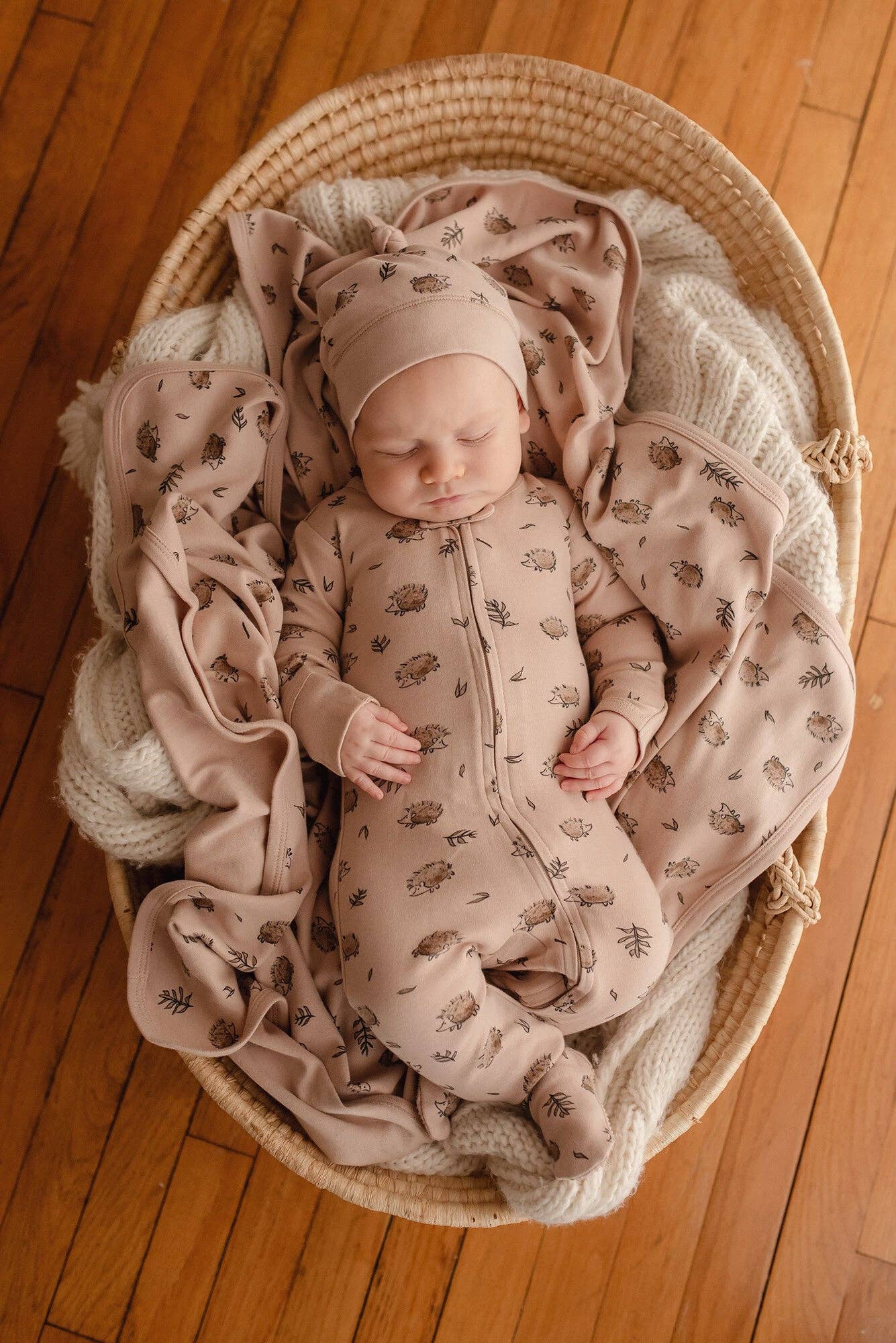 Newborn baby in a crib with beige hedgehog print bedding and matching footie and haton a wooden floor