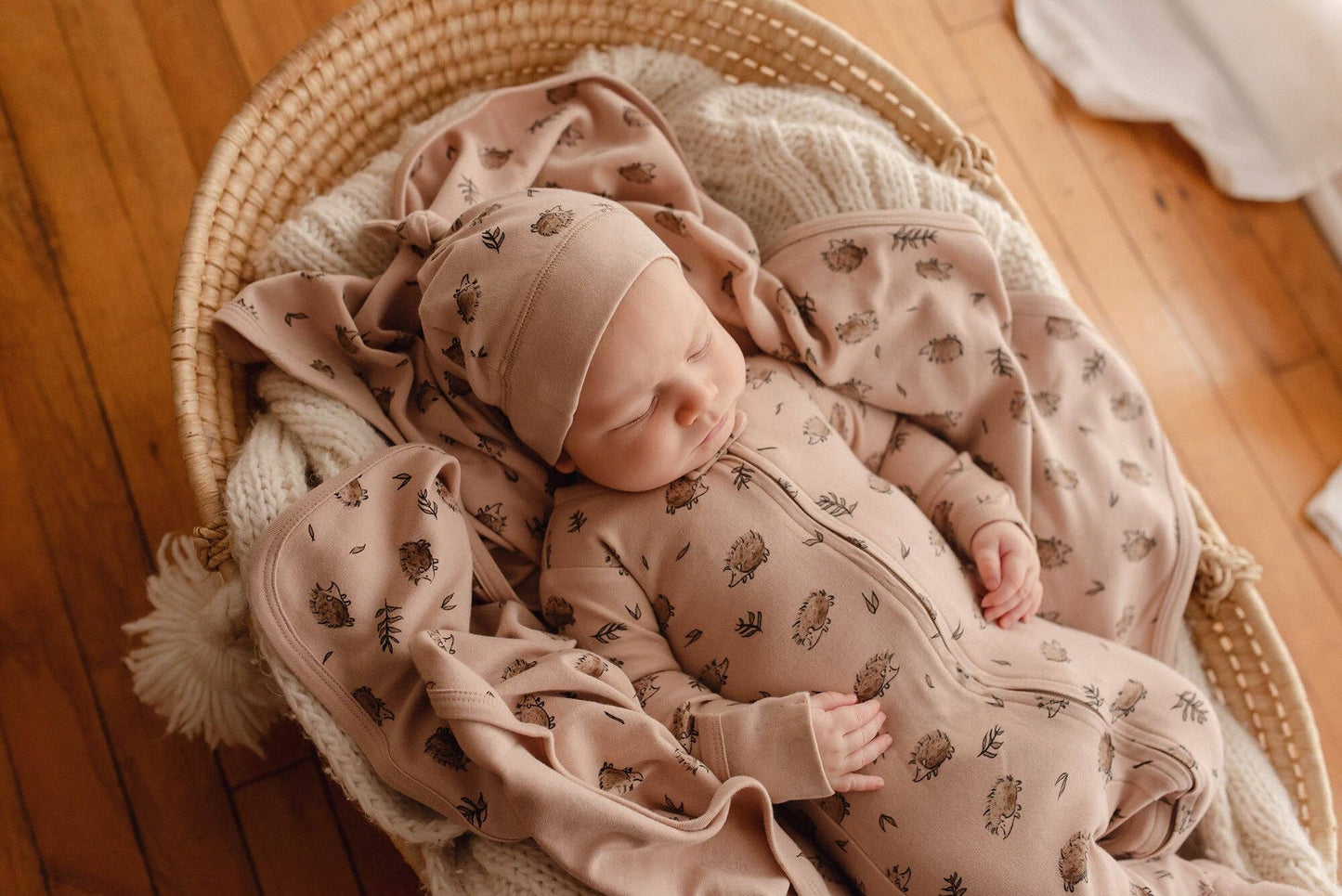 Newborn baby in a crib with matching beige hedgehog romper, blanket and hat on a wooden floor