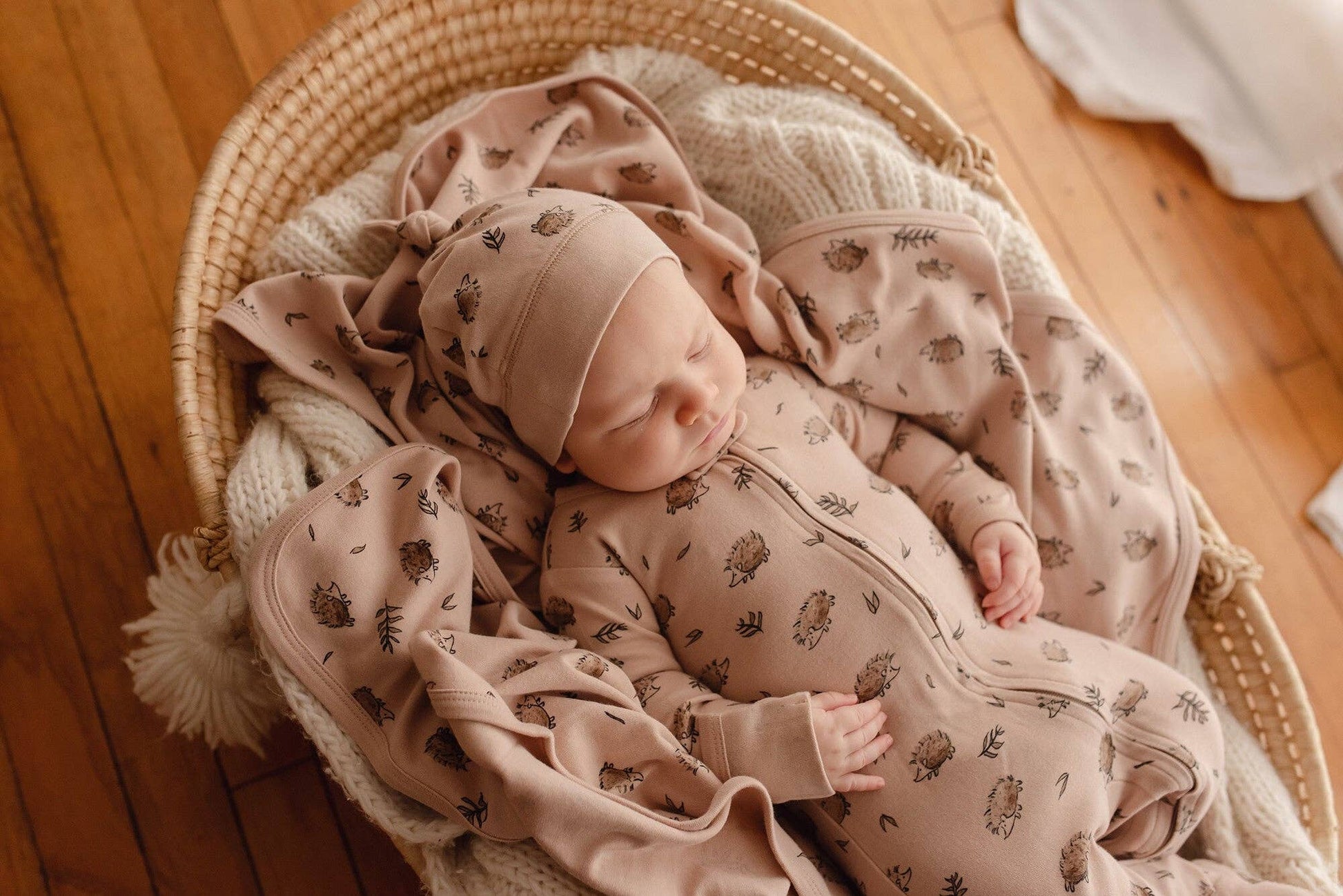 Newborn baby in a crib with matching beige hedgehog romper, blanket and hat on a wooden floor