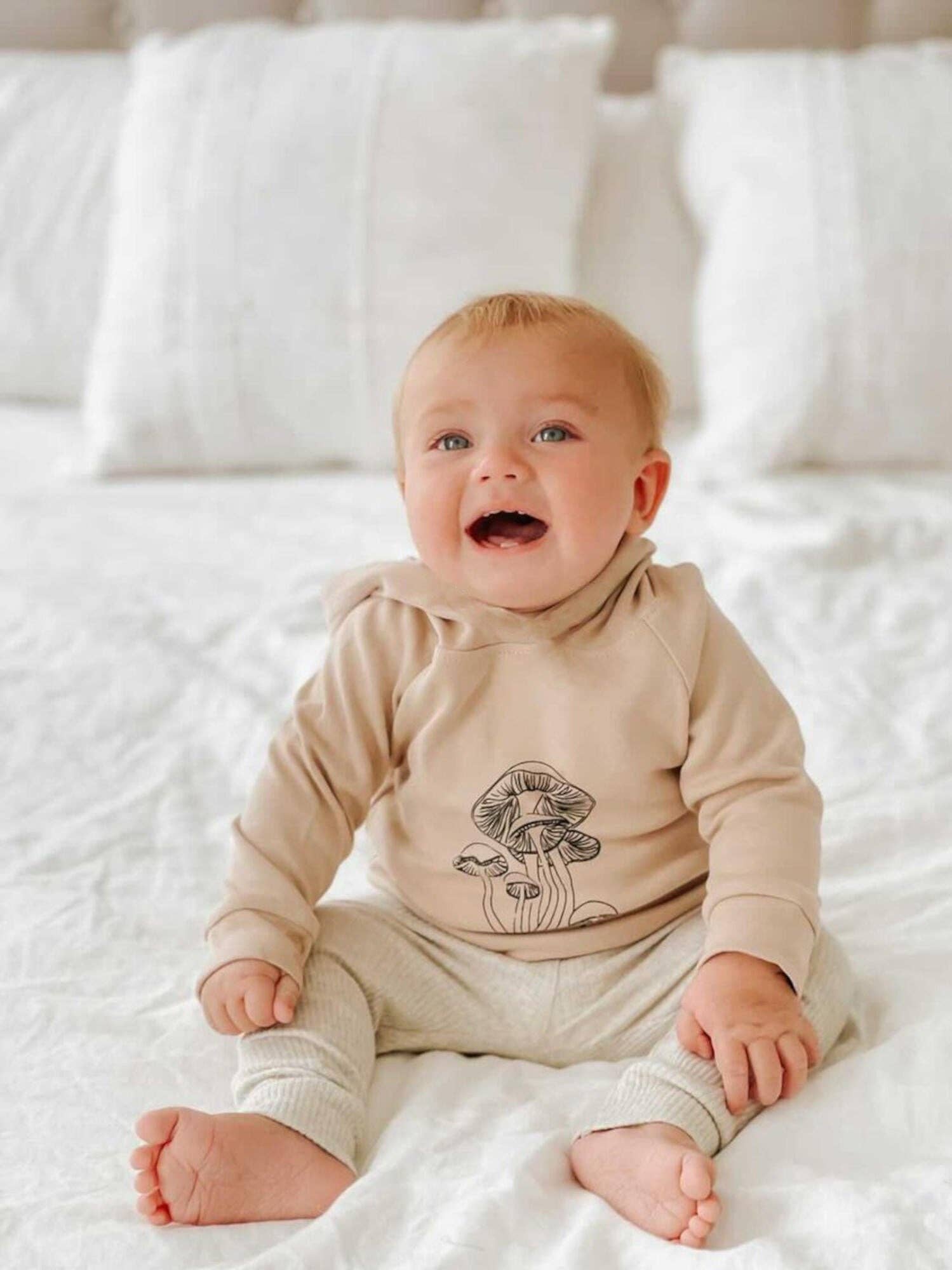 Baby wearing a beige hoodie with a mushroom design on it sitting on a white bed